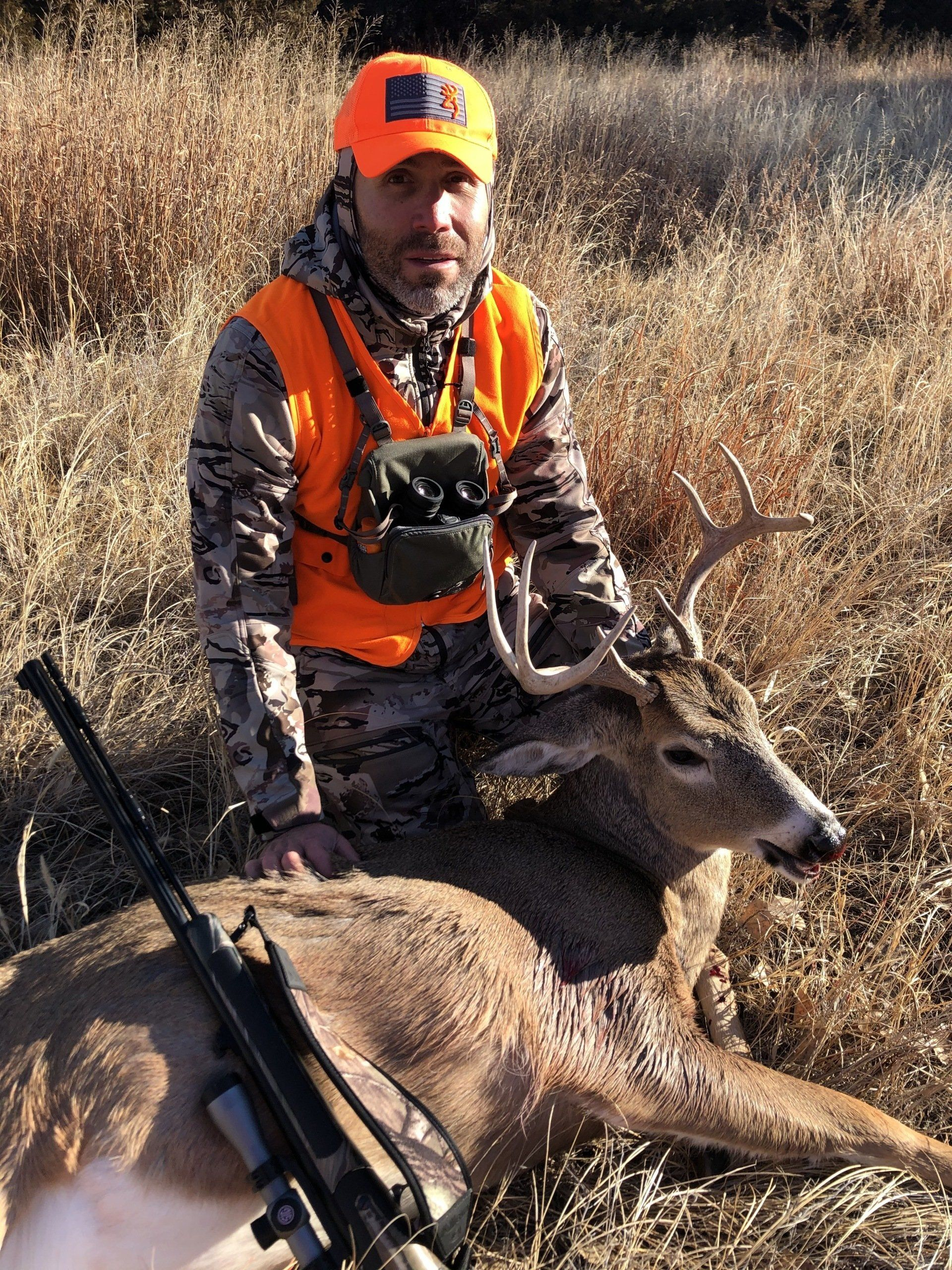 Hunter in orange vest kneeling beside a deer in tall dry grass, holding a rifle.