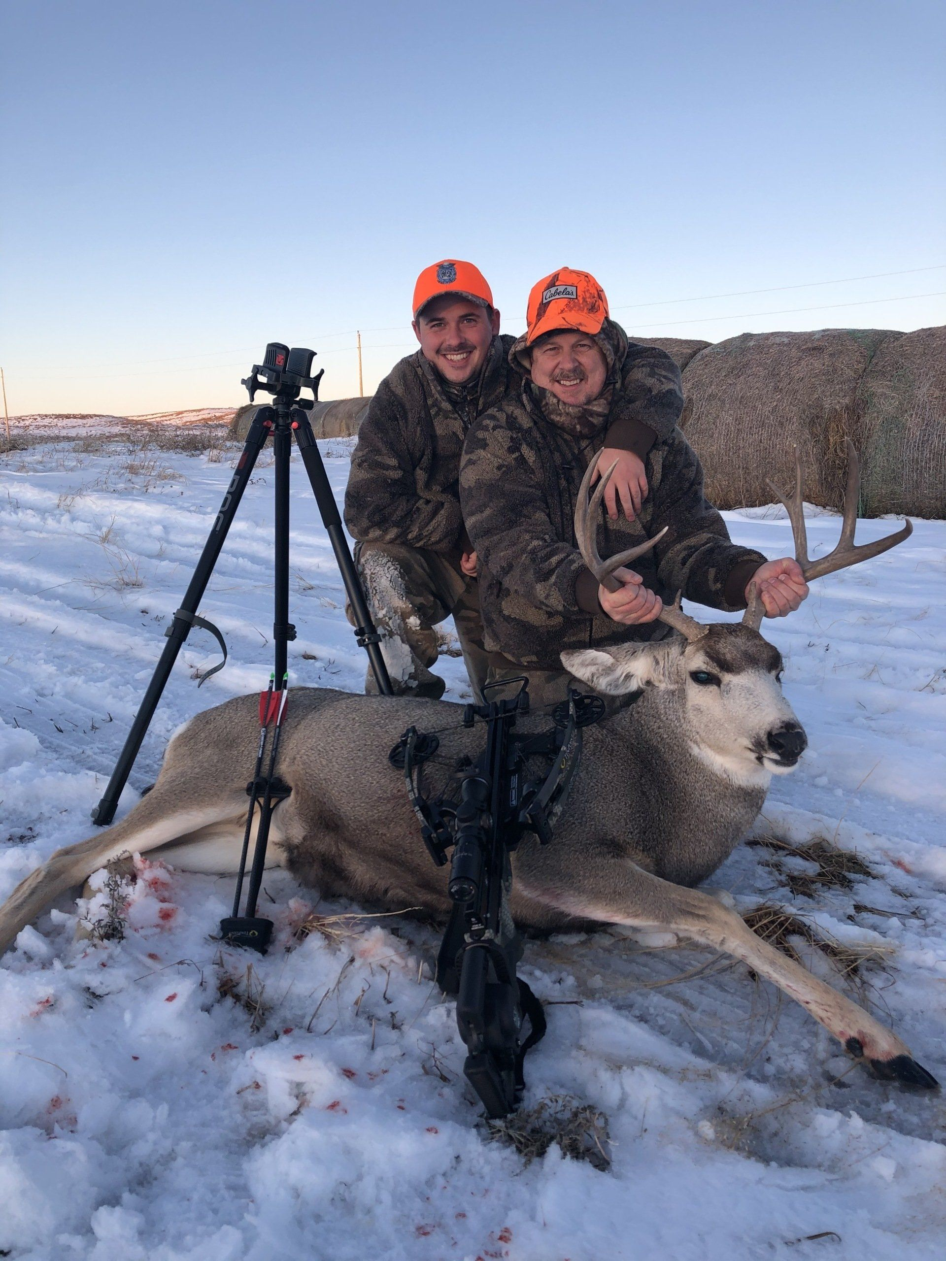 Two hunters pose with a deer in snowy field beside a tripod at sunset