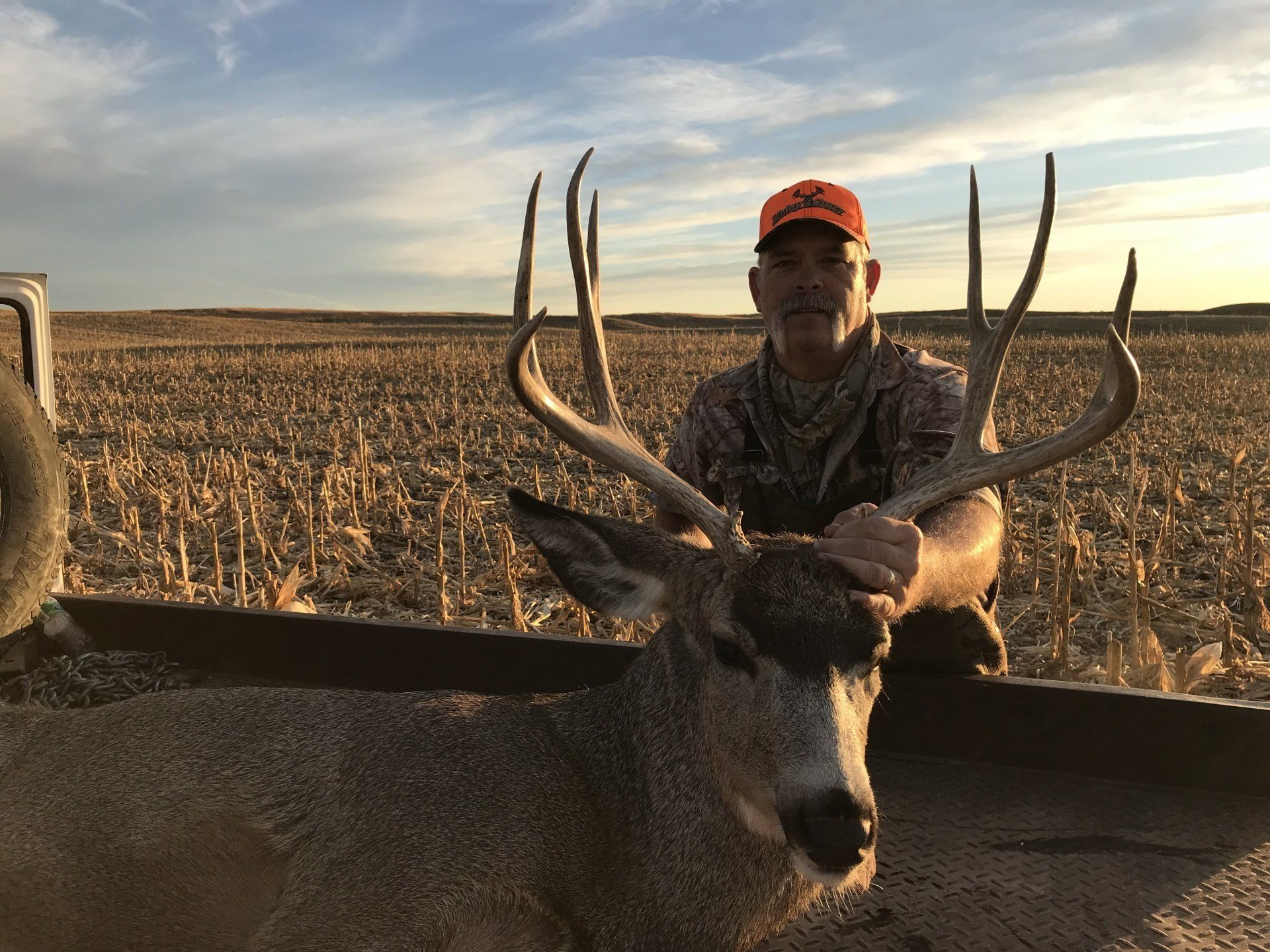 Hunter posing with a large buck in a harvested field at sunset.