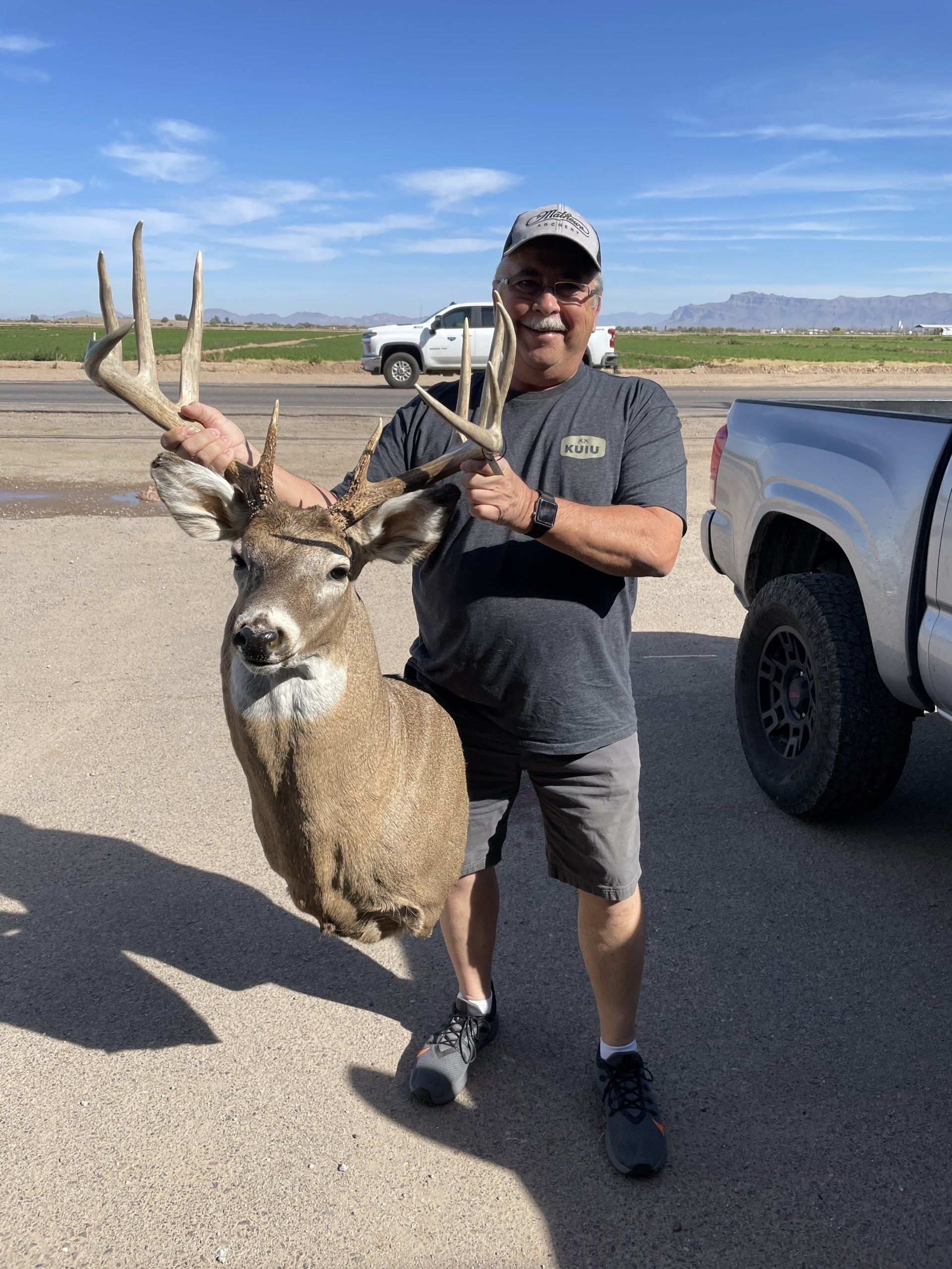 Hunter holding a large deer by the antlers beside a pickup truck on a sunny roadside.