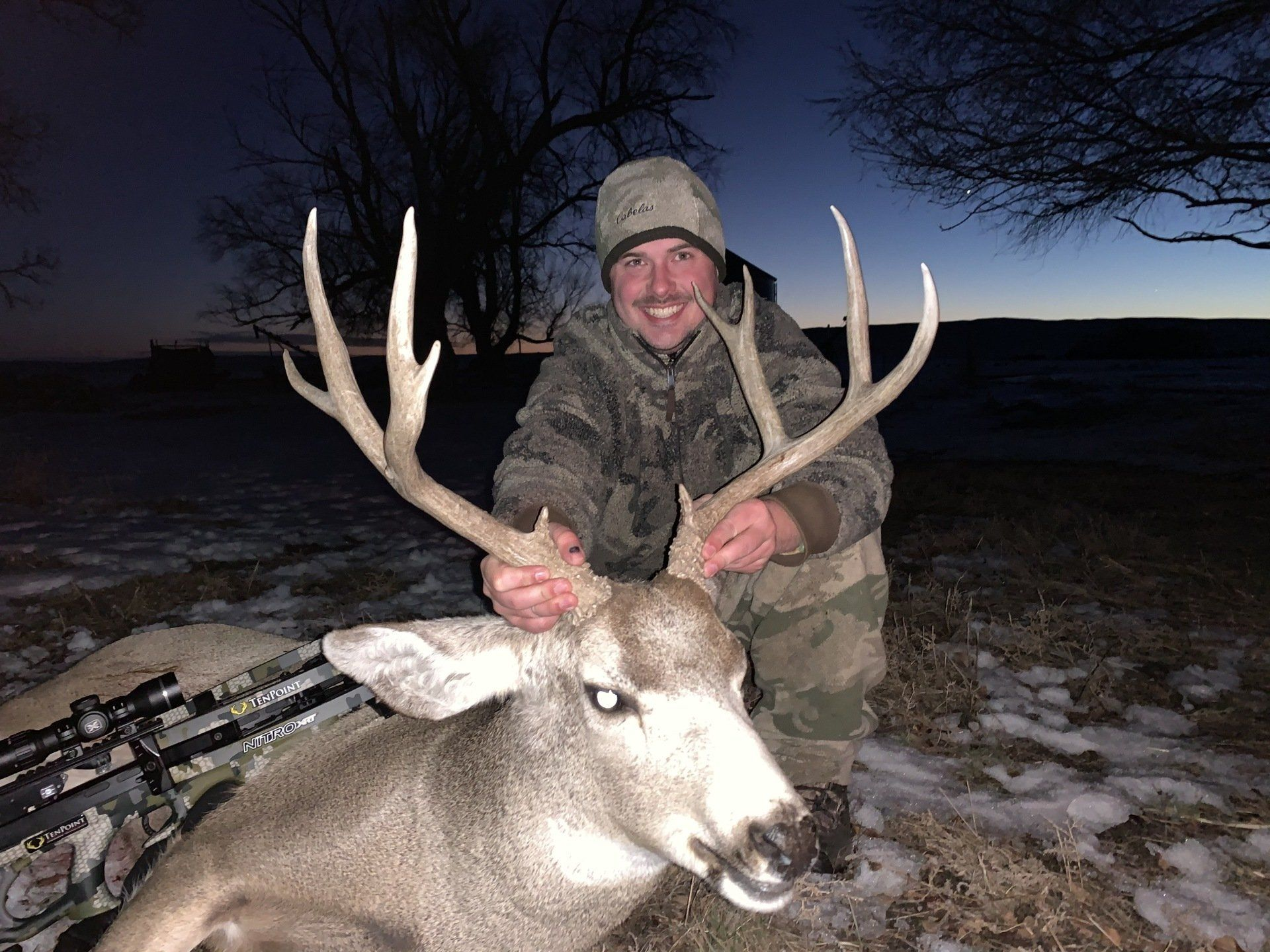 Hunter posing with a large buck at dusk in a snowy field