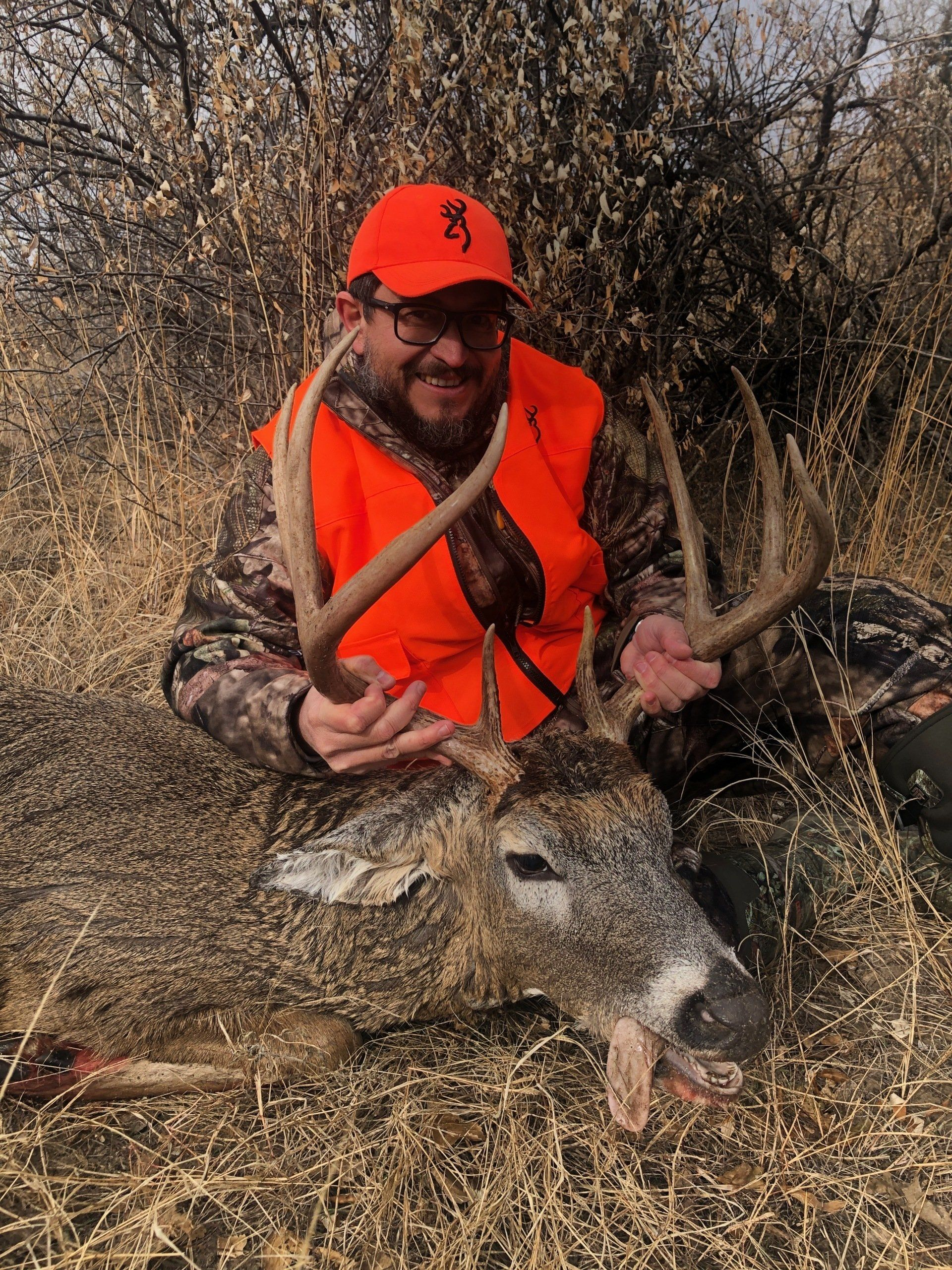 Hunter in orange vest and cap holding a deer in dry brush.