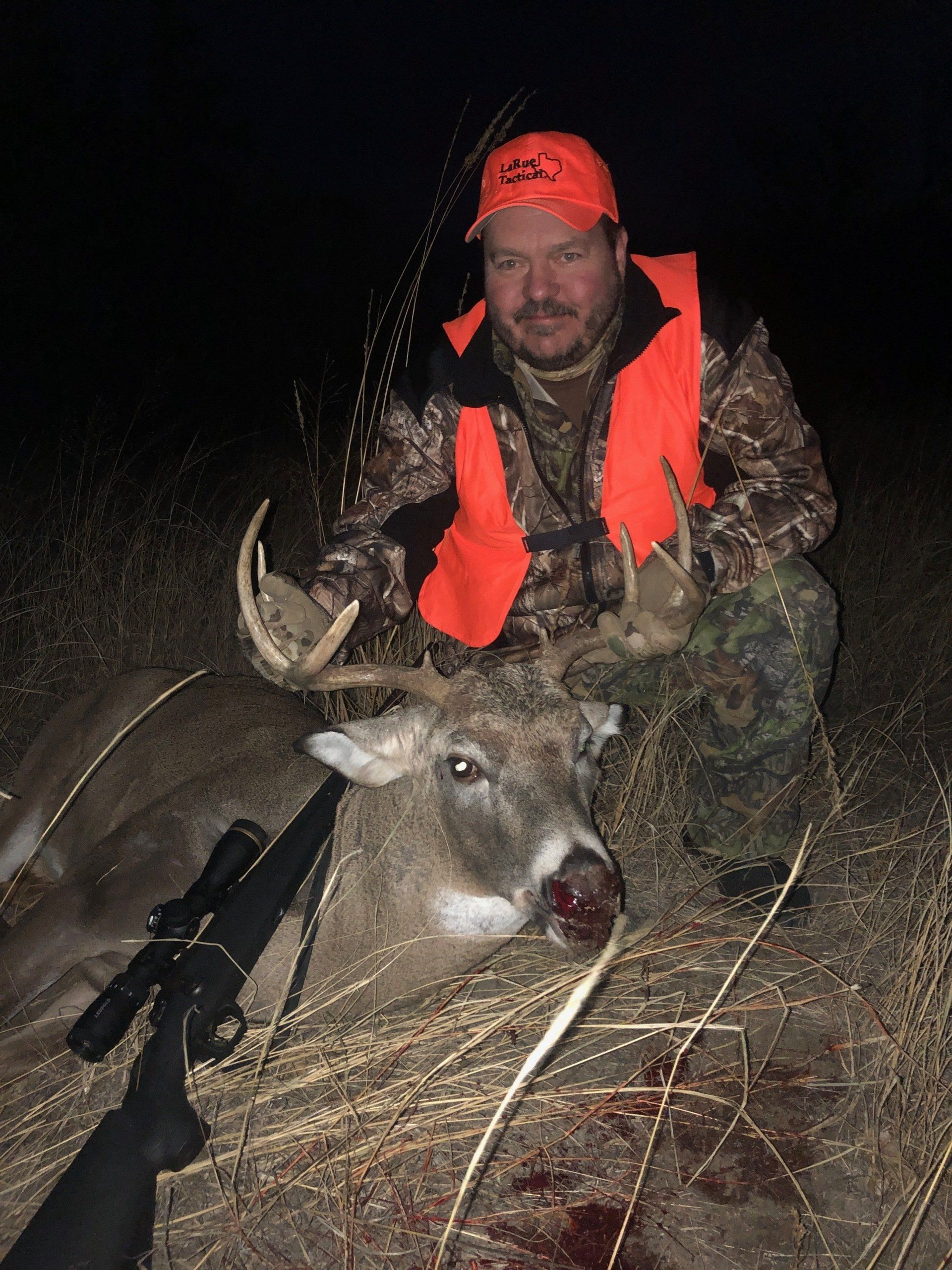 Hunter in orange vest and cap kneels beside a deer he has hunted at night.
