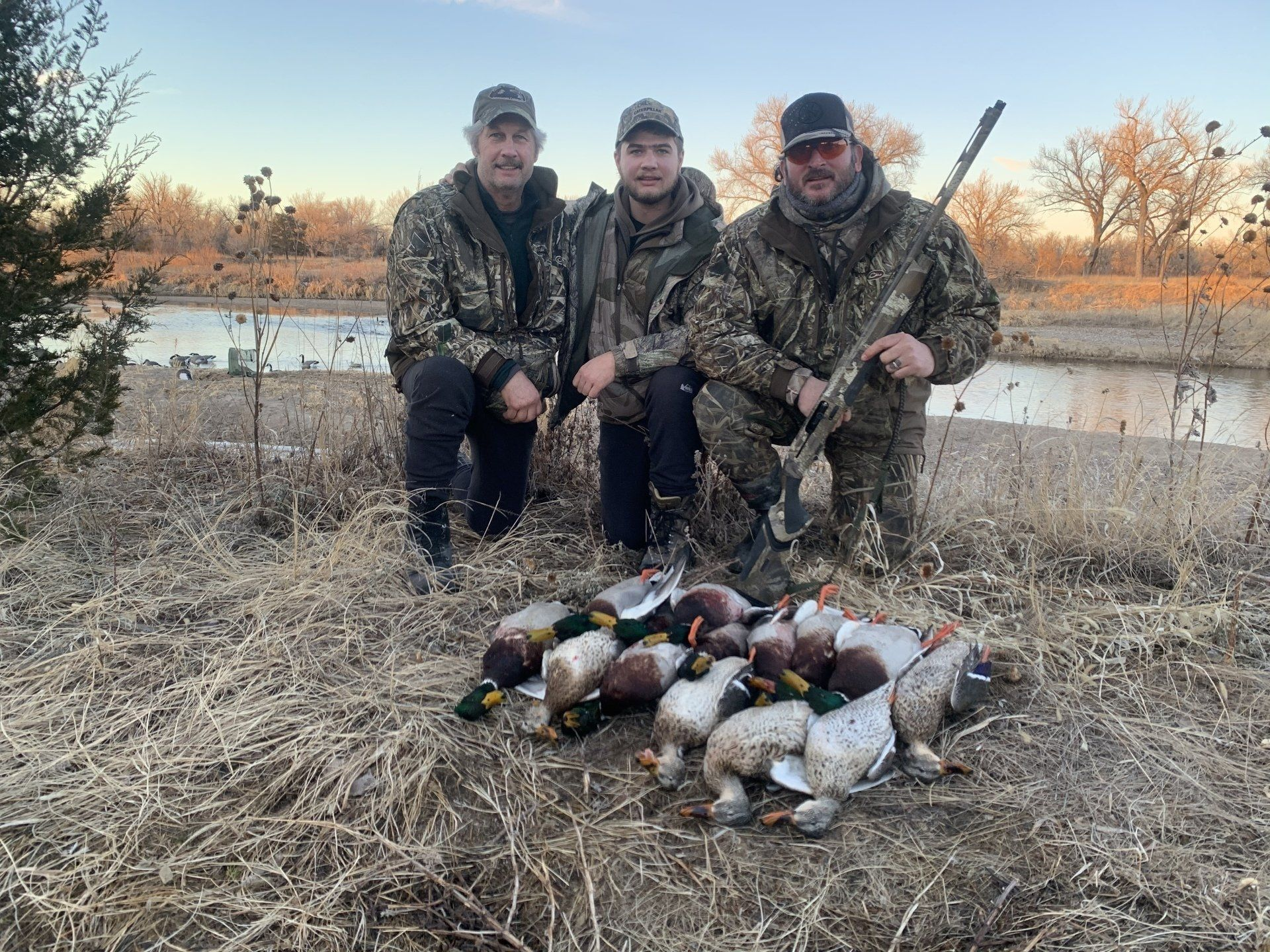 Three hunters in camouflage pose kneeling beside a pile of ducks by a riverside at sunset.