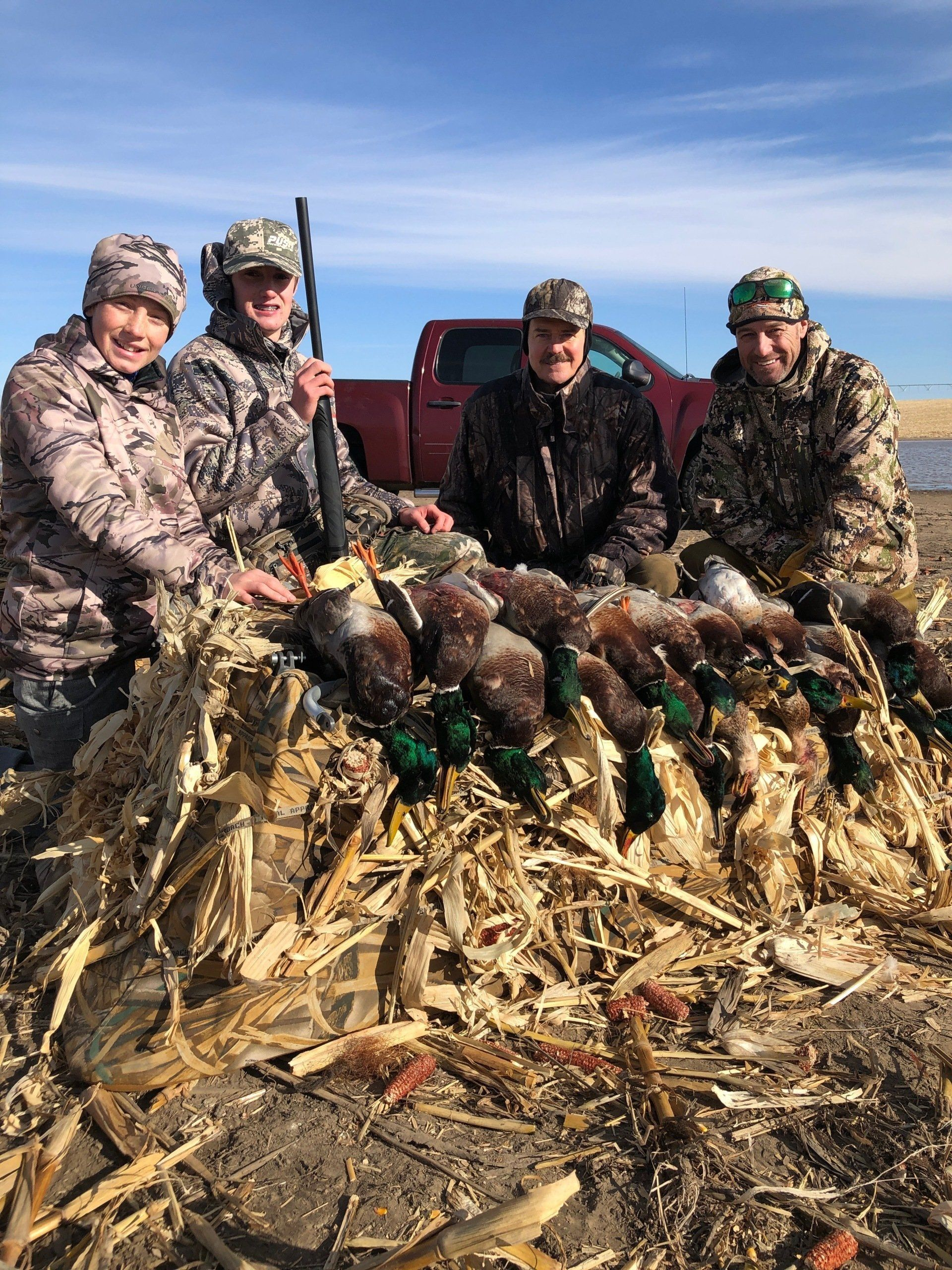 Four hunters pose behind a pile of harvested ducks in a snowy field under a clear sky.
