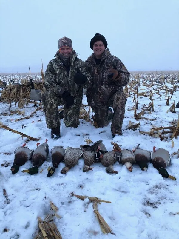 Two hunters in camo kneel in a snowy cornfield behind a row of harvested geese.