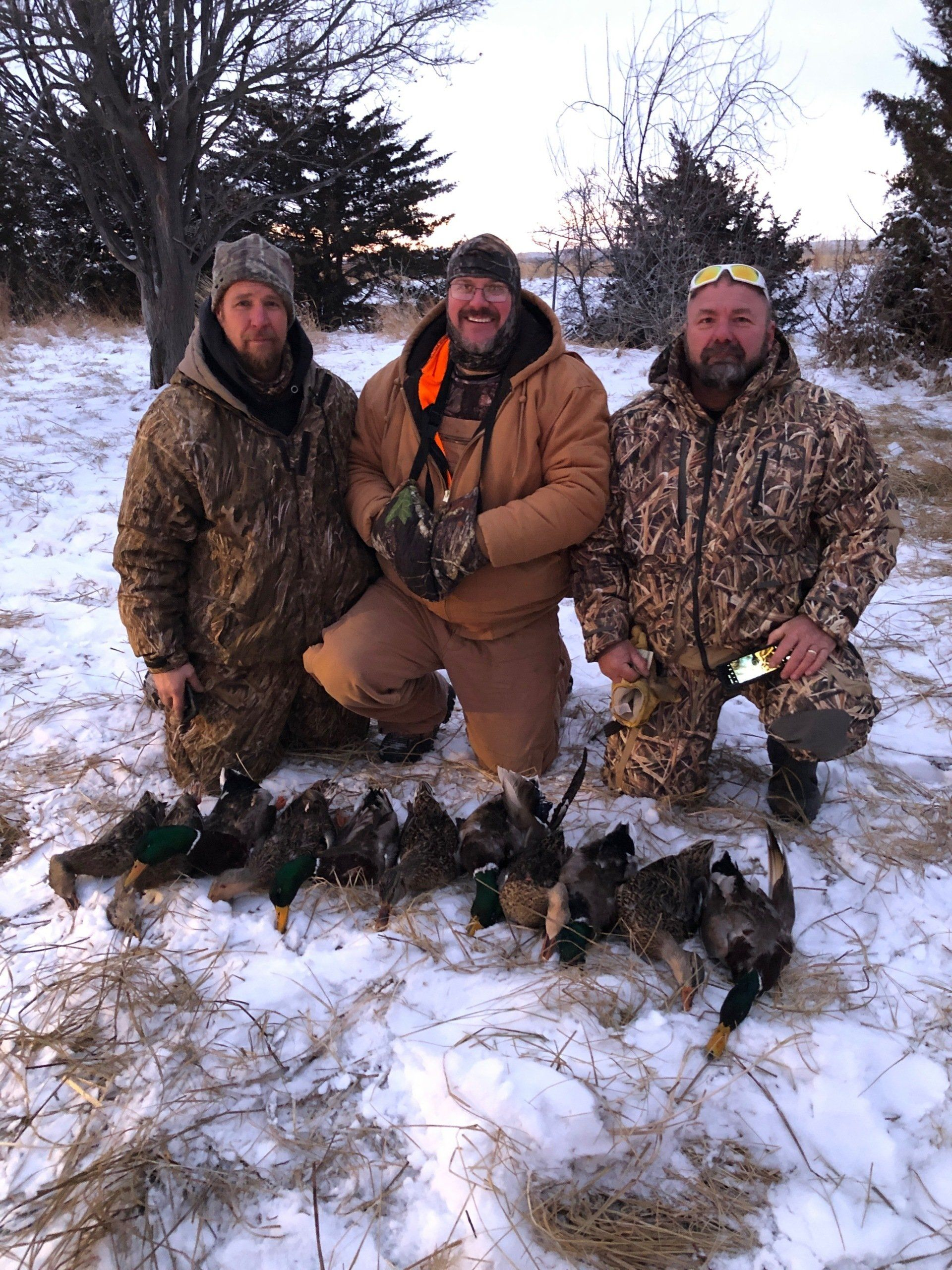 Three hunters in camouflage kneel in snow beside a row of ducks at dusk