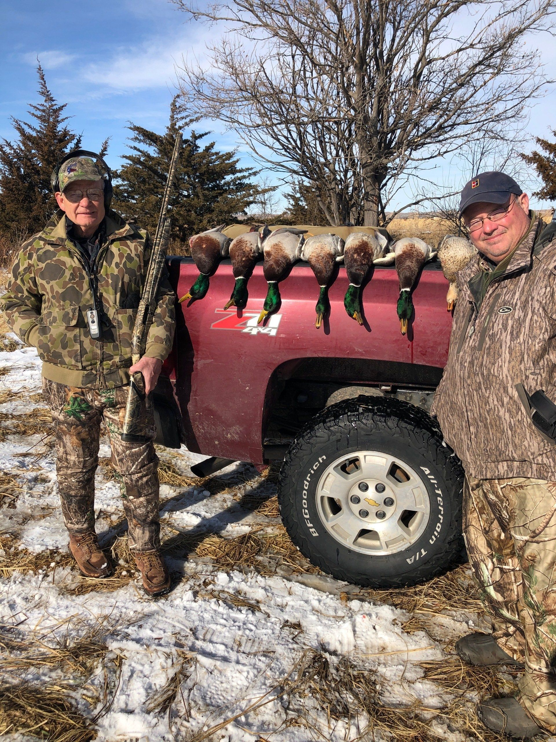 Two people in camouflage stand beside a red pickup truck on a snowy field with deer hanging from the side.