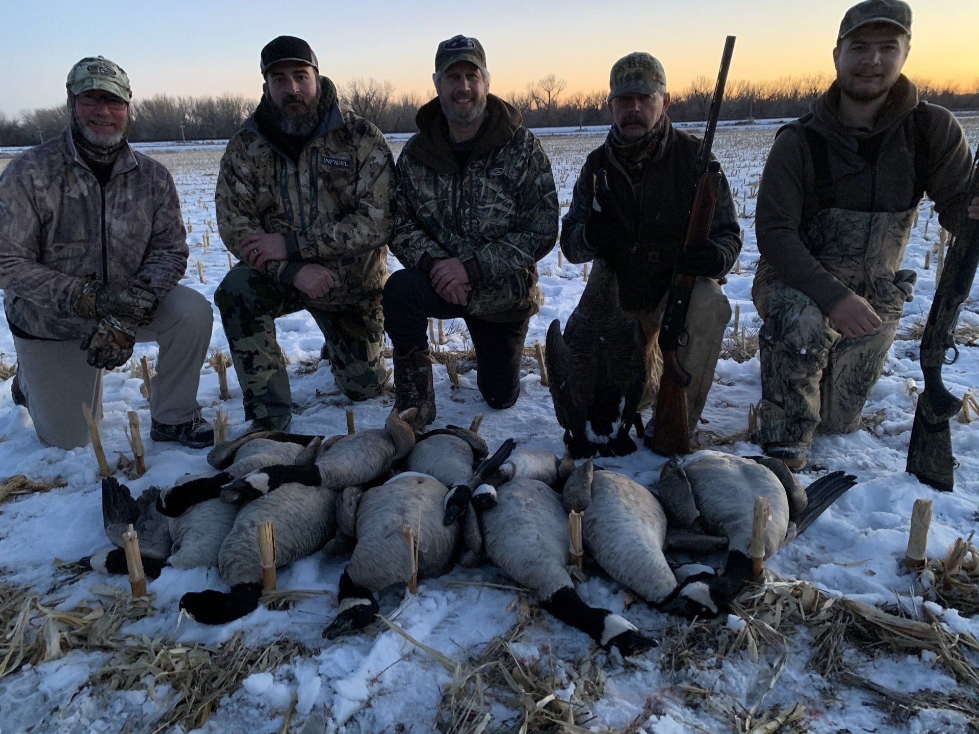 Five hunters in camouflage pose behind a large pile of harvested geese in a snowy field at sunset