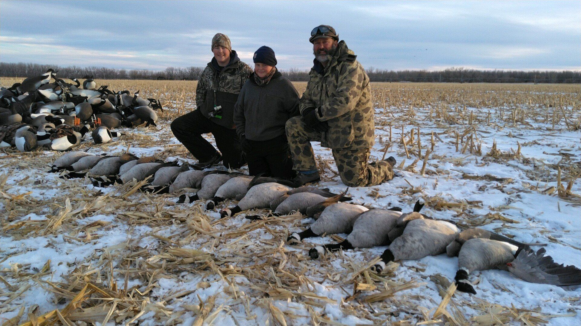 Three people kneel beside a row of large fish on snowy ground in a field, with a pile of fish at left.