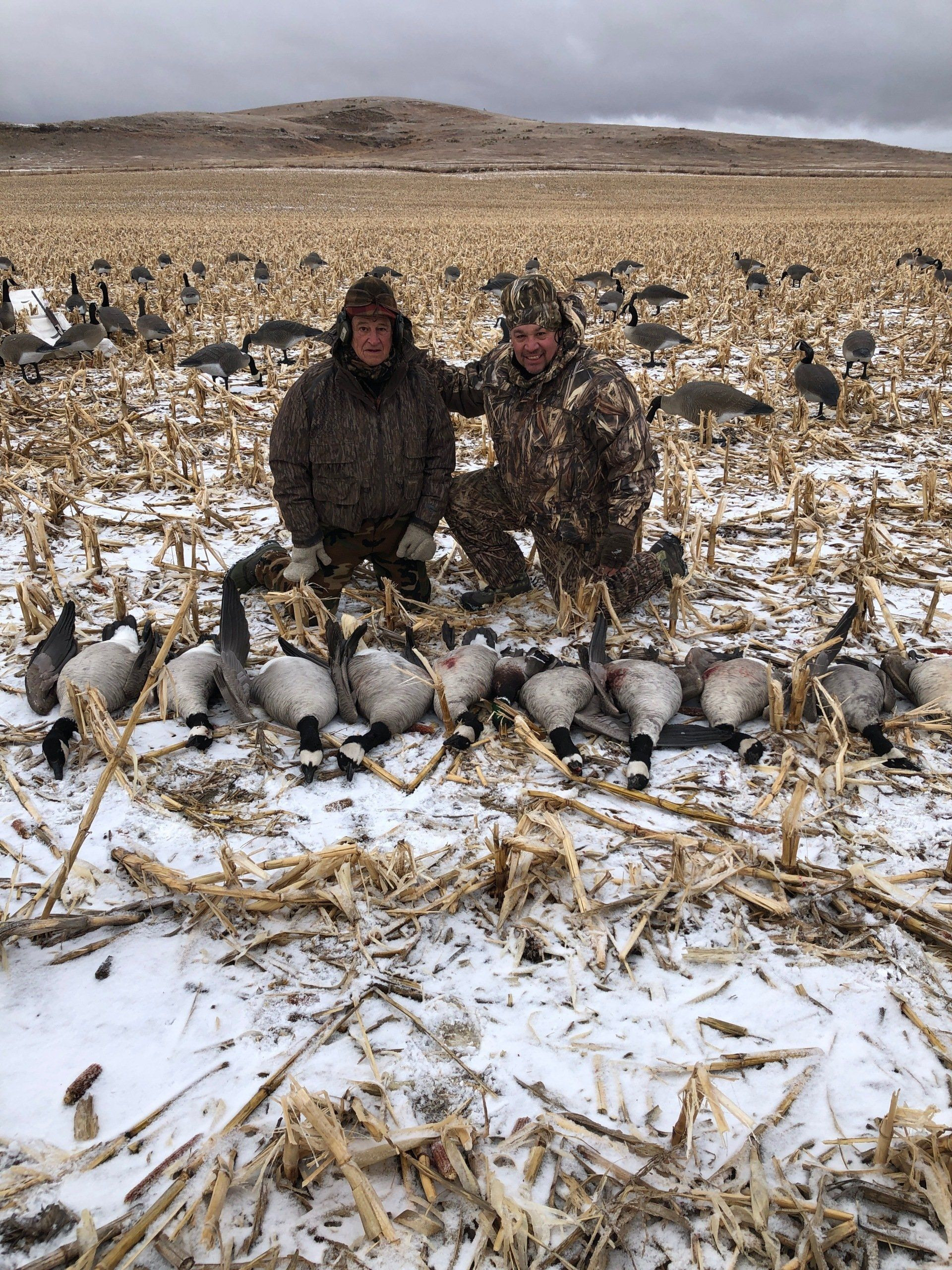 Two hunters kneel in a snowy field beside a large pile of harvested birds.