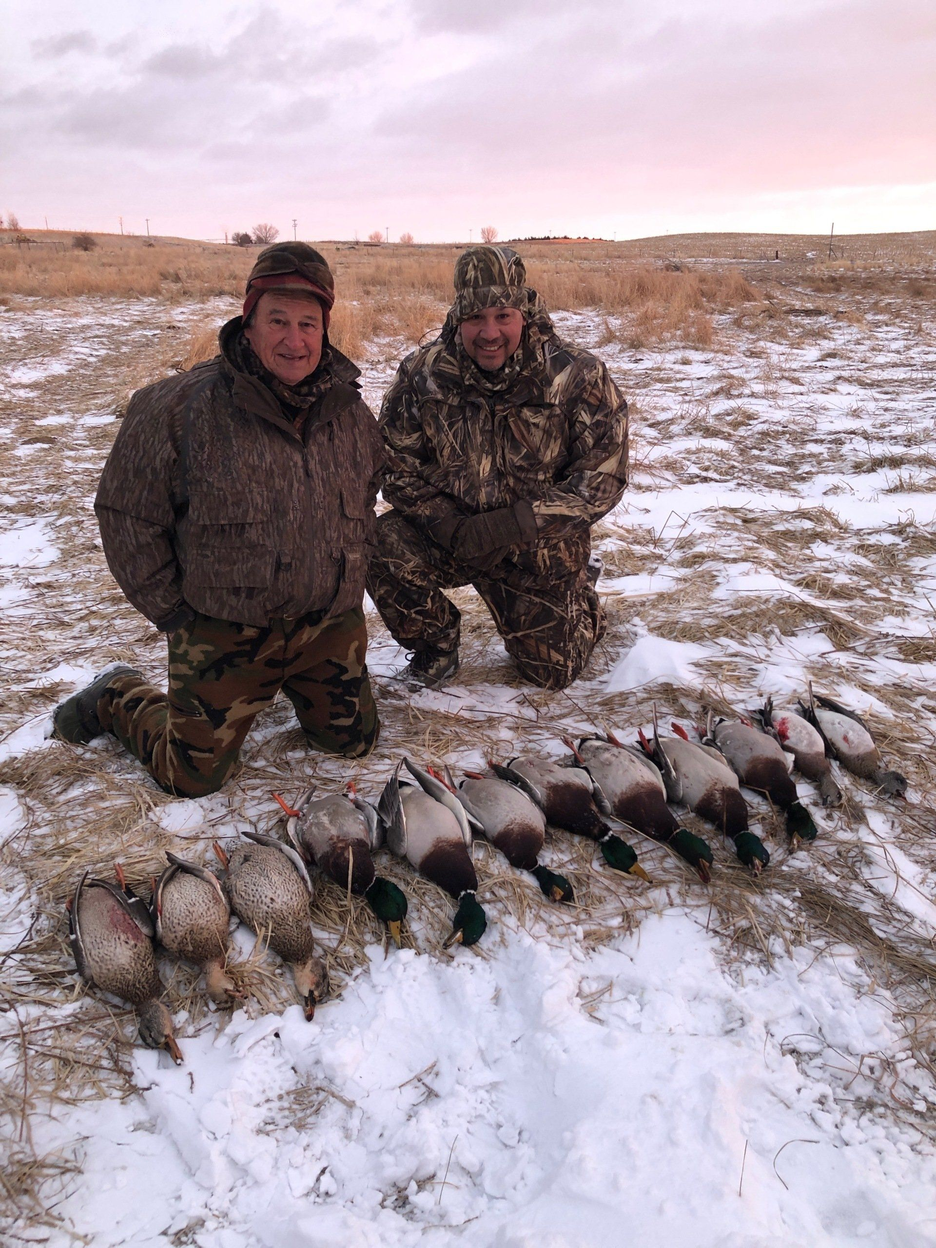 Two people kneel in snowy field beside a row of caught fish and ducks.