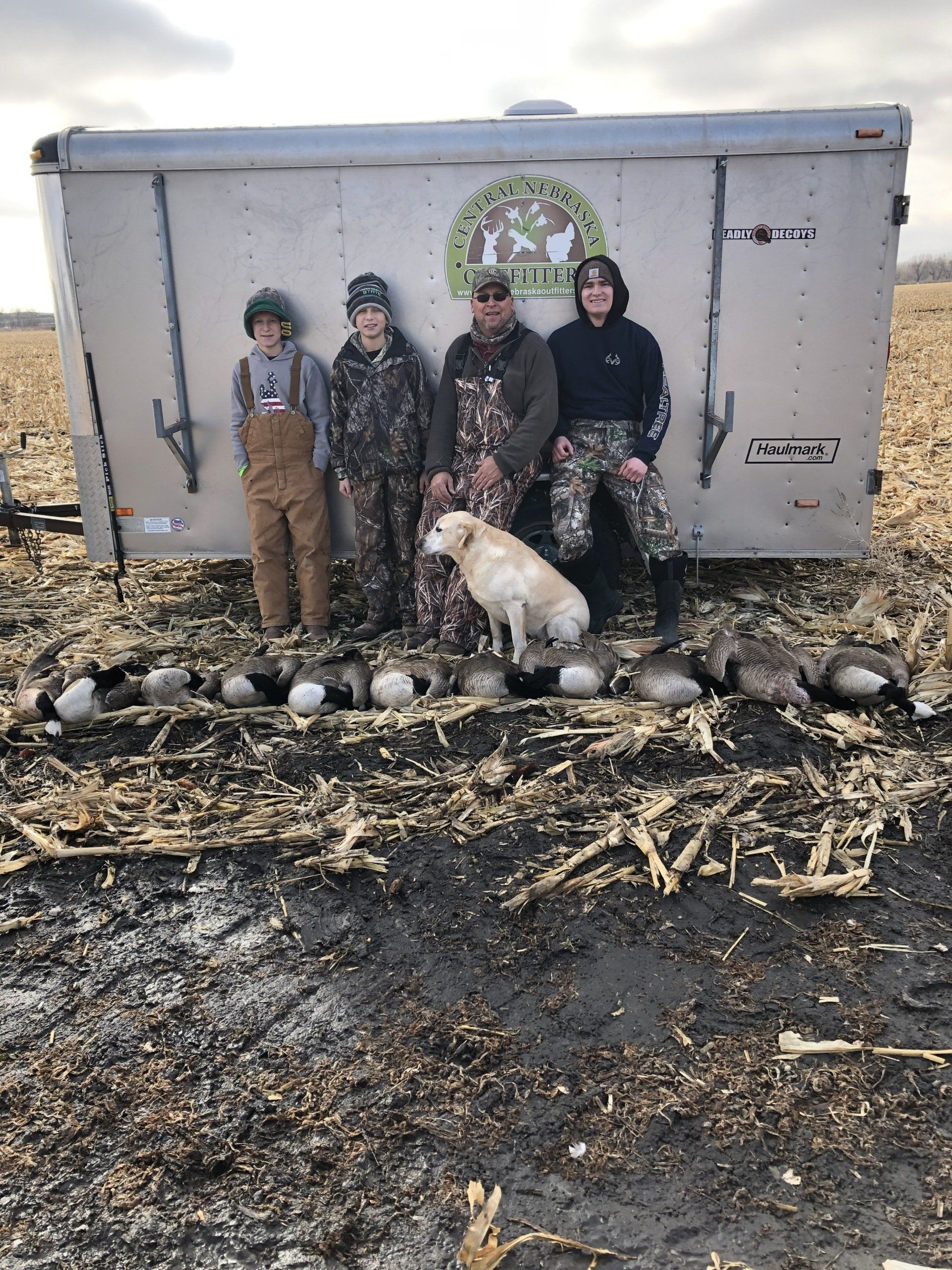 Four people and a white dog posing outdoors in front of a gray trailer on a muddy lot.