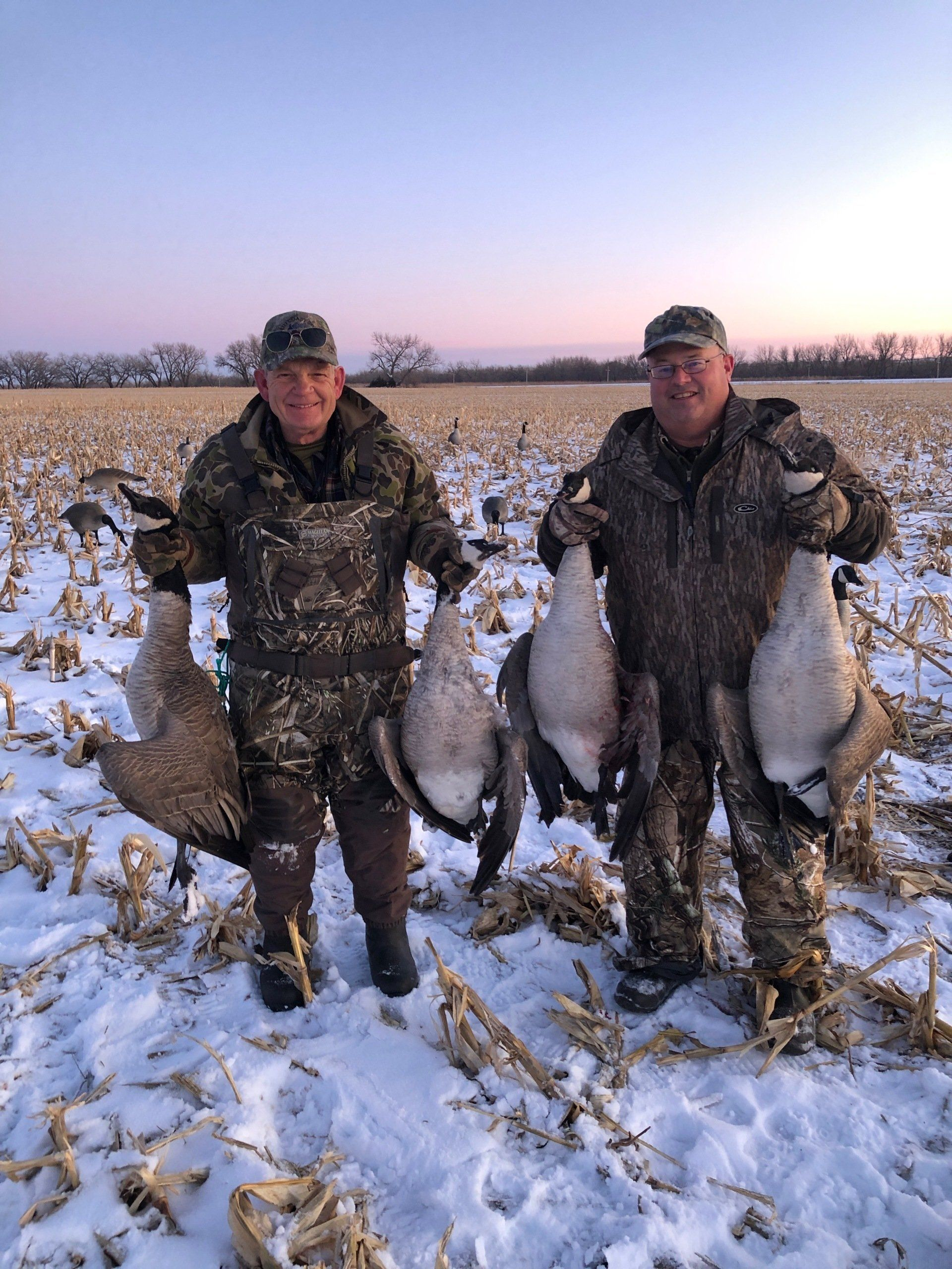 Two hunters in snowy field holding four large ducks at dusk