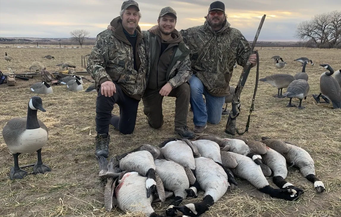 Three hunters pose with a pile of geese in a field, with more geese standing behind them.