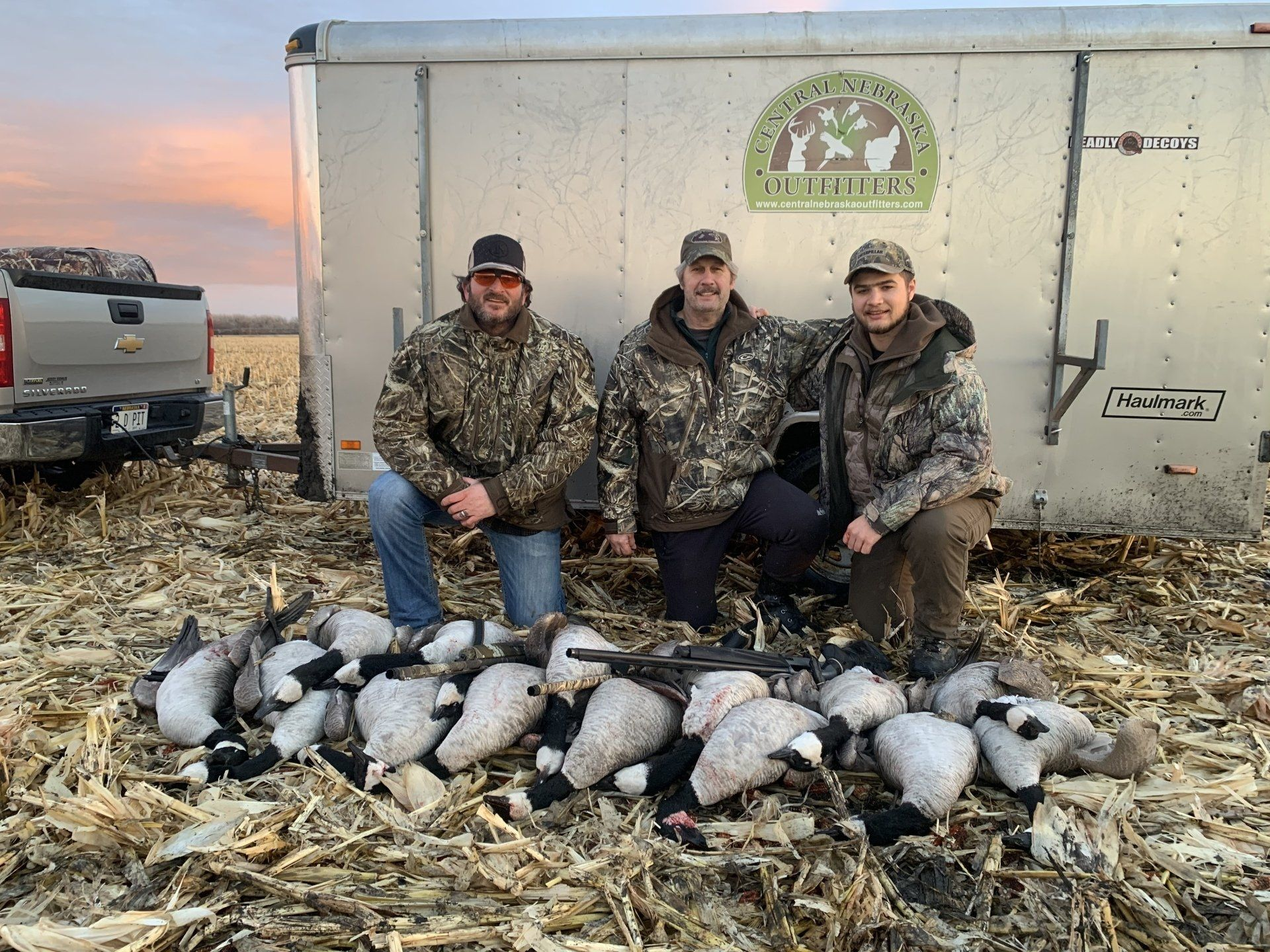 Three hunters kneel behind a row of harvested ducks in a field beside a trailer.