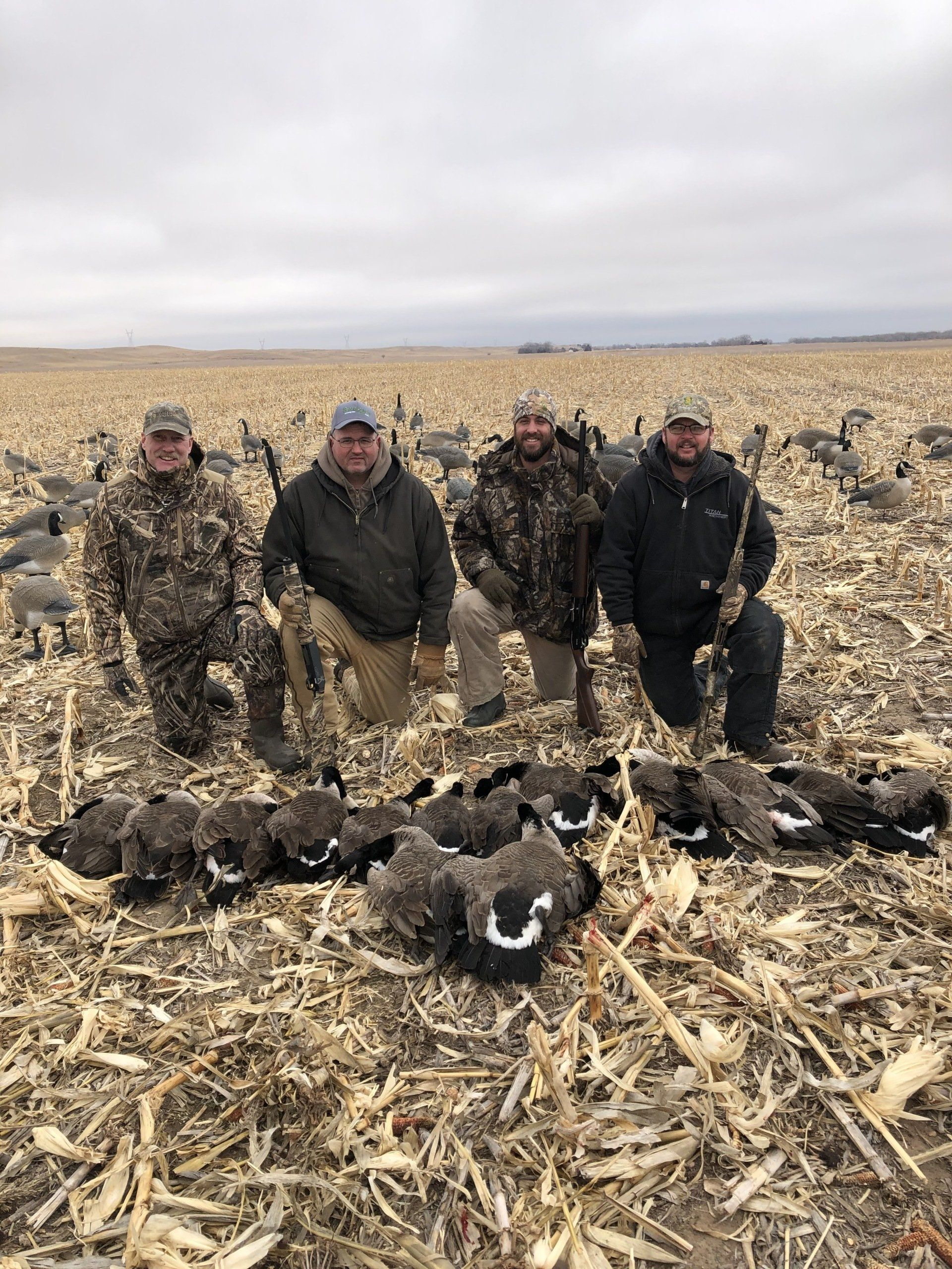 Four hunters stand in a harvested field behind a pile of downed ducks under an overcast sky.