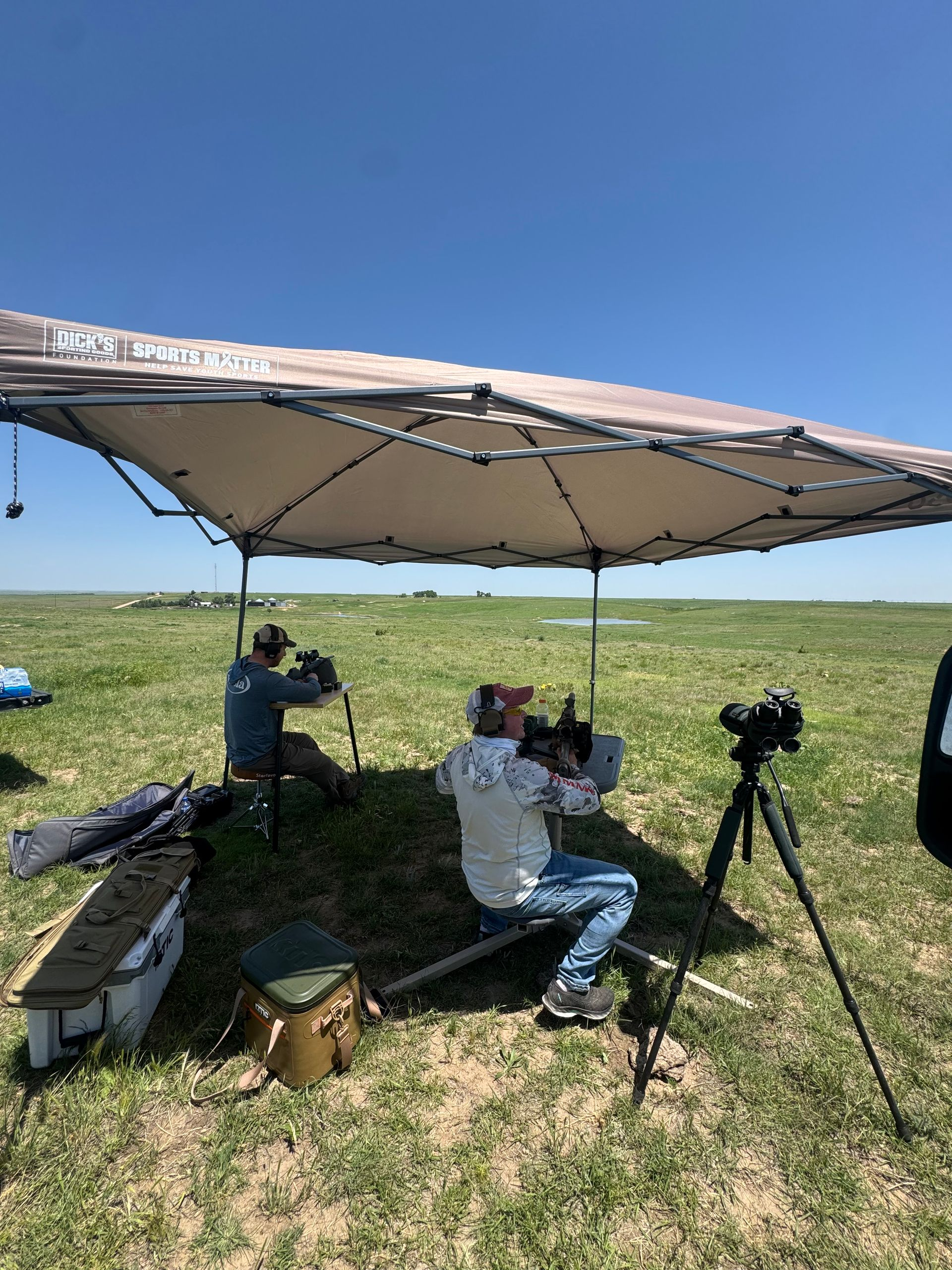 People seated under a sunshade canopy in a grassy field, with a tripod nearby.