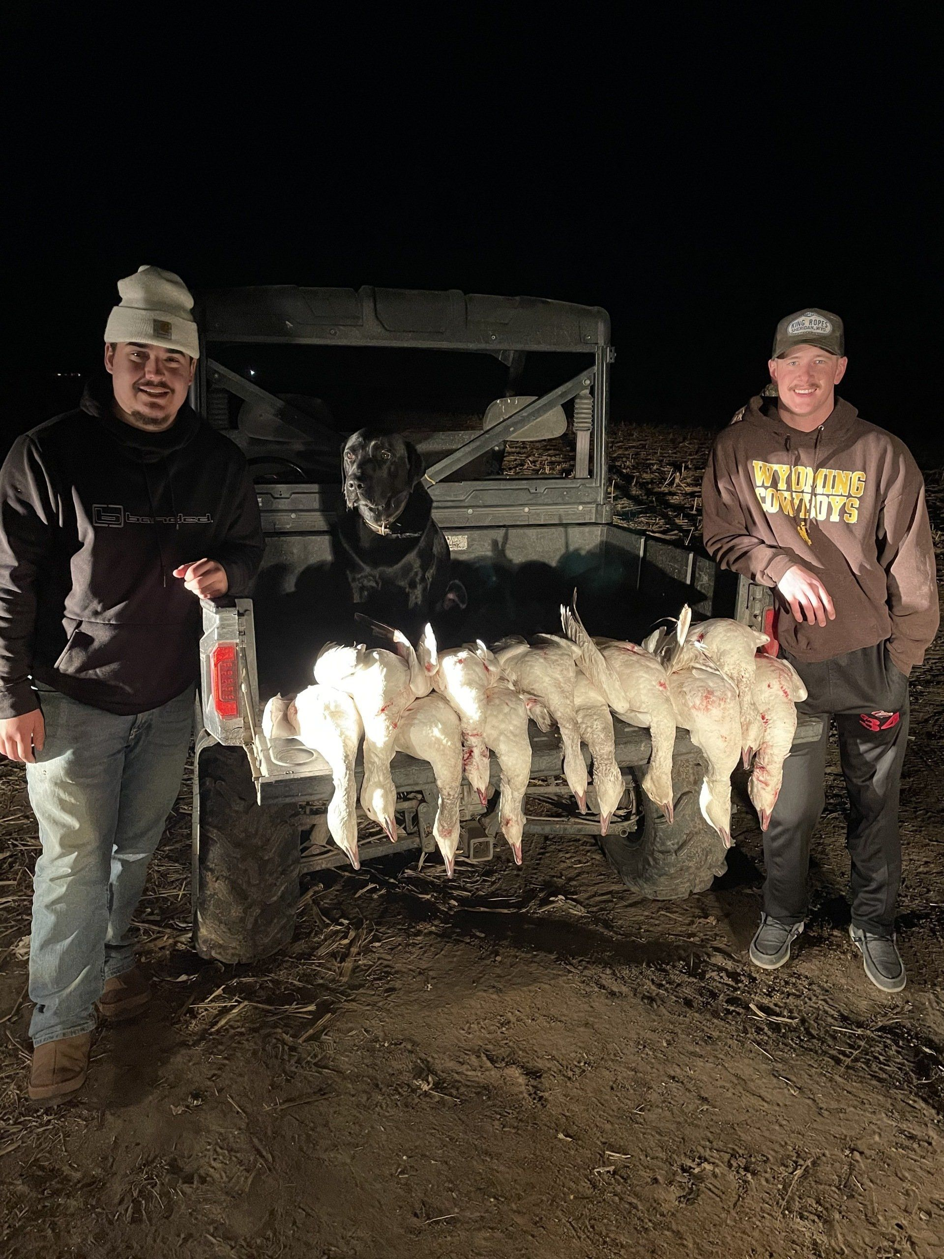 Two people pose at night with a row of hunted birds hanging from a rack near a truck.
