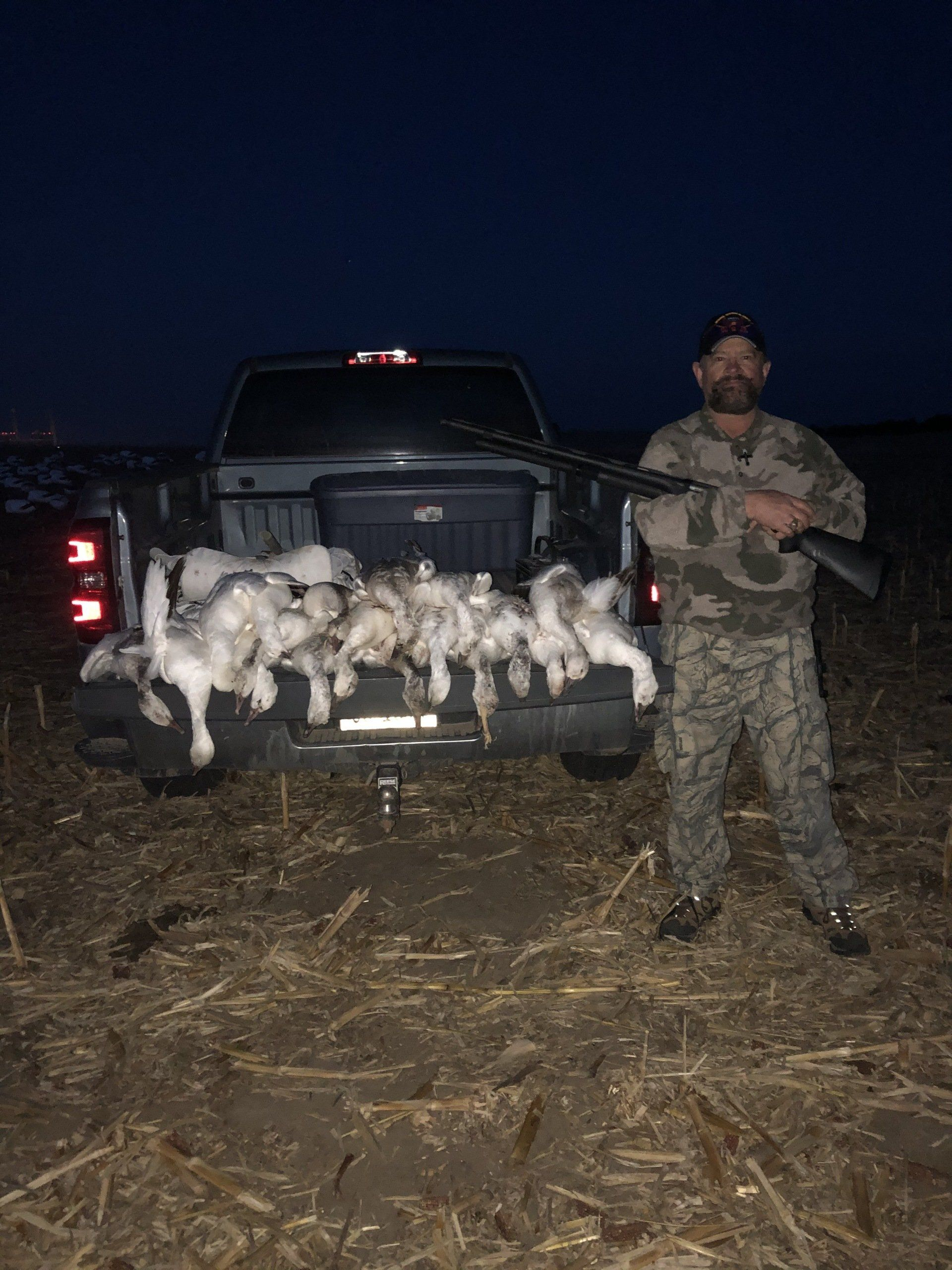 Nighttime hunting scene with a man holding a rifle beside a truck bed full of hunted geese.