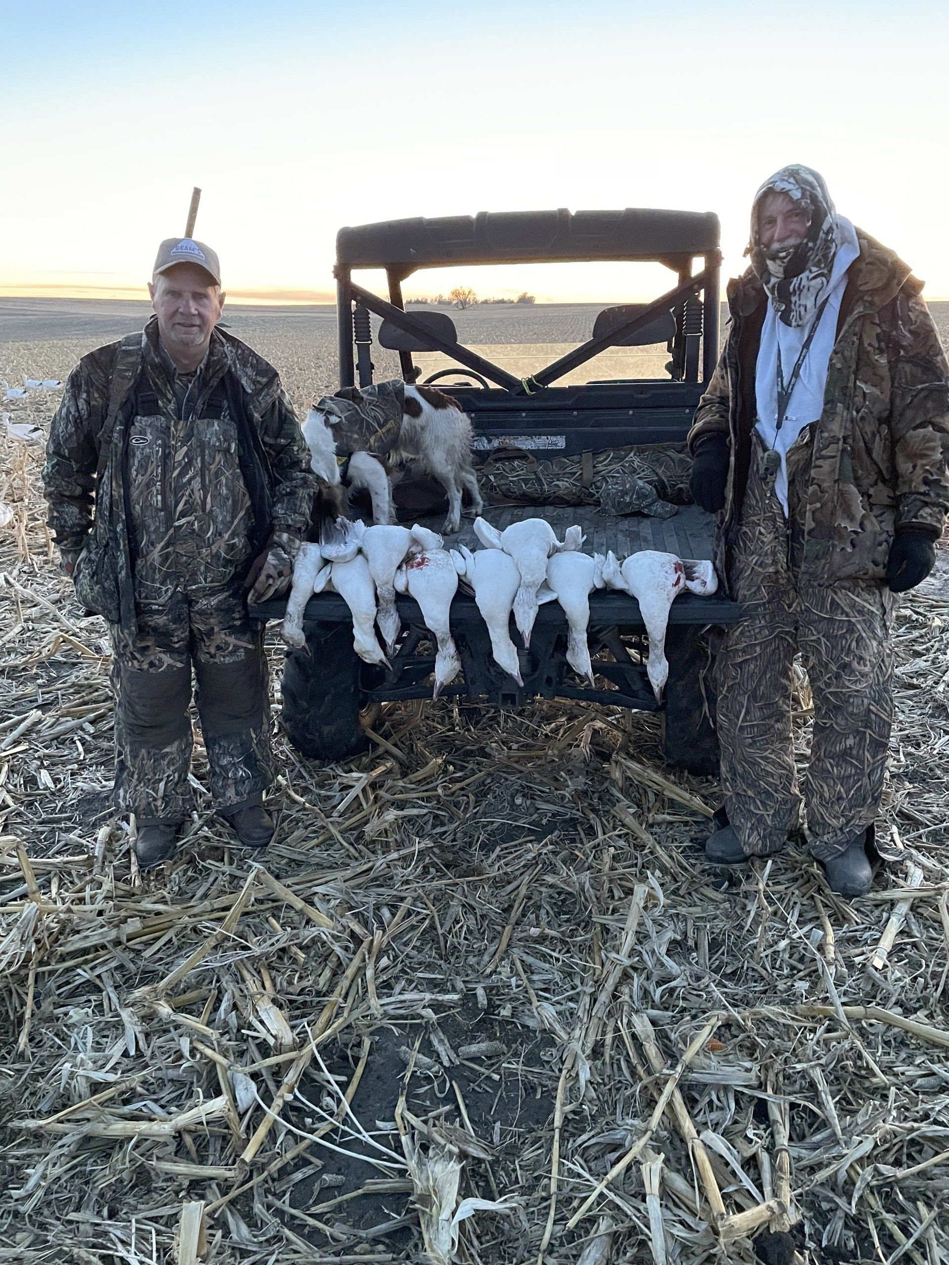Two hunters standing by a UTV with several harvested coyotes on the front rack in a snowy field.