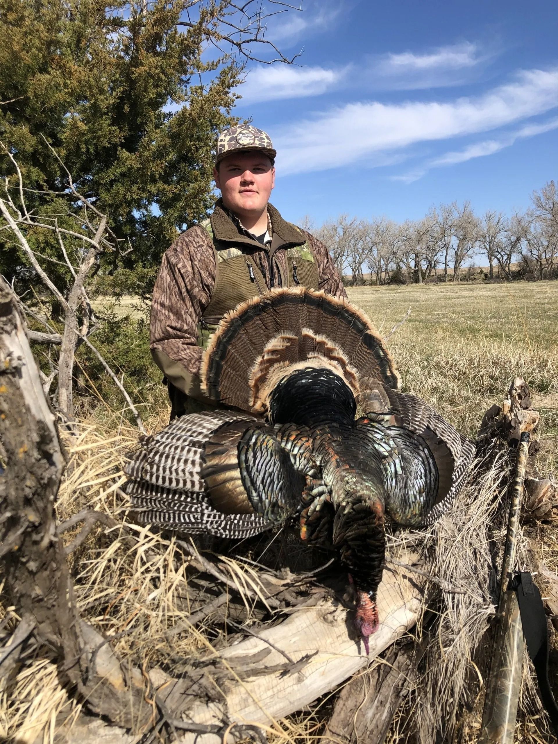 Hunter posing outdoors with a turkey in brush under a blue sky