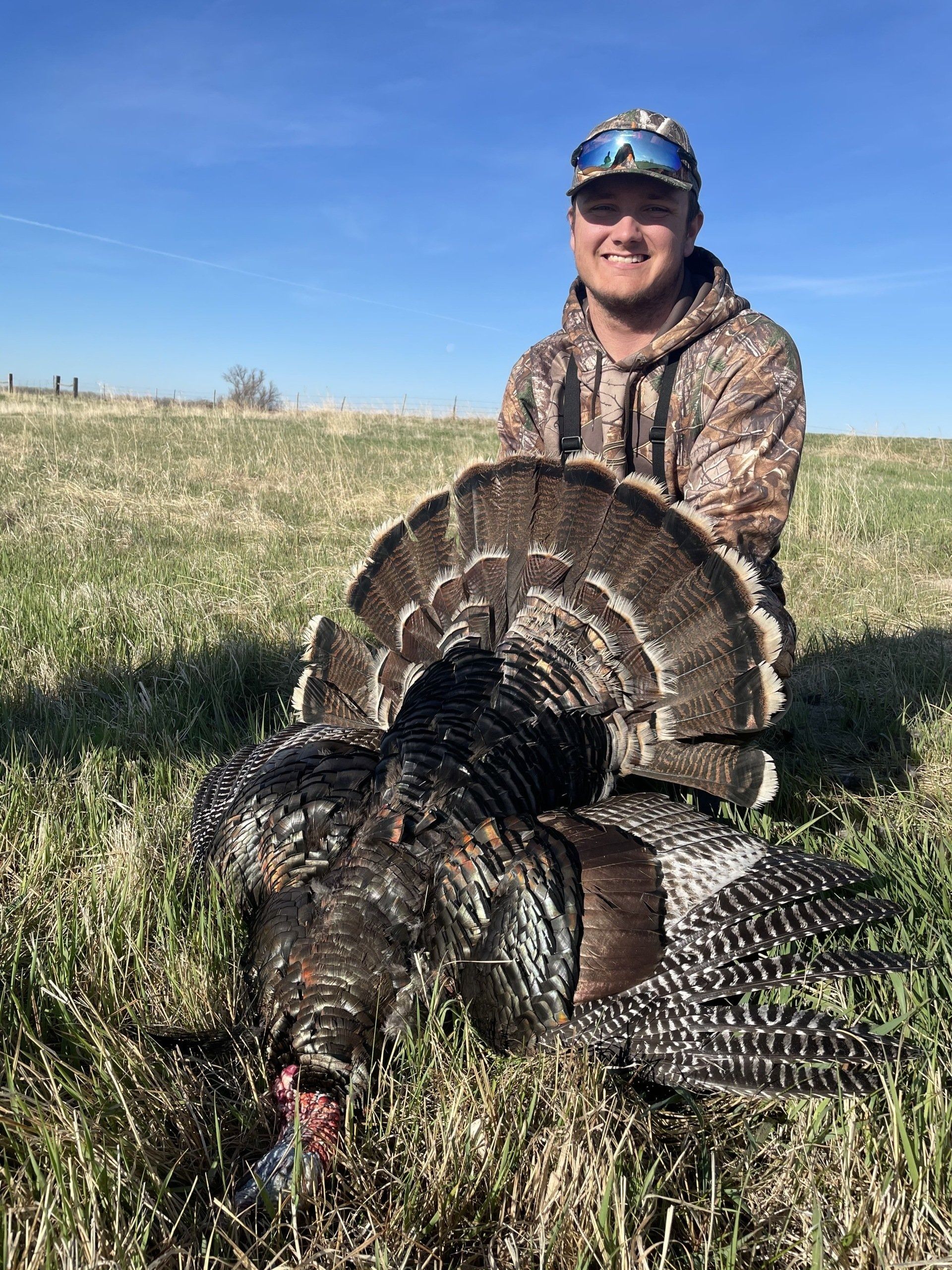 Hunter kneeling beside a harvested wild turkey in a grassy field under a blue sky