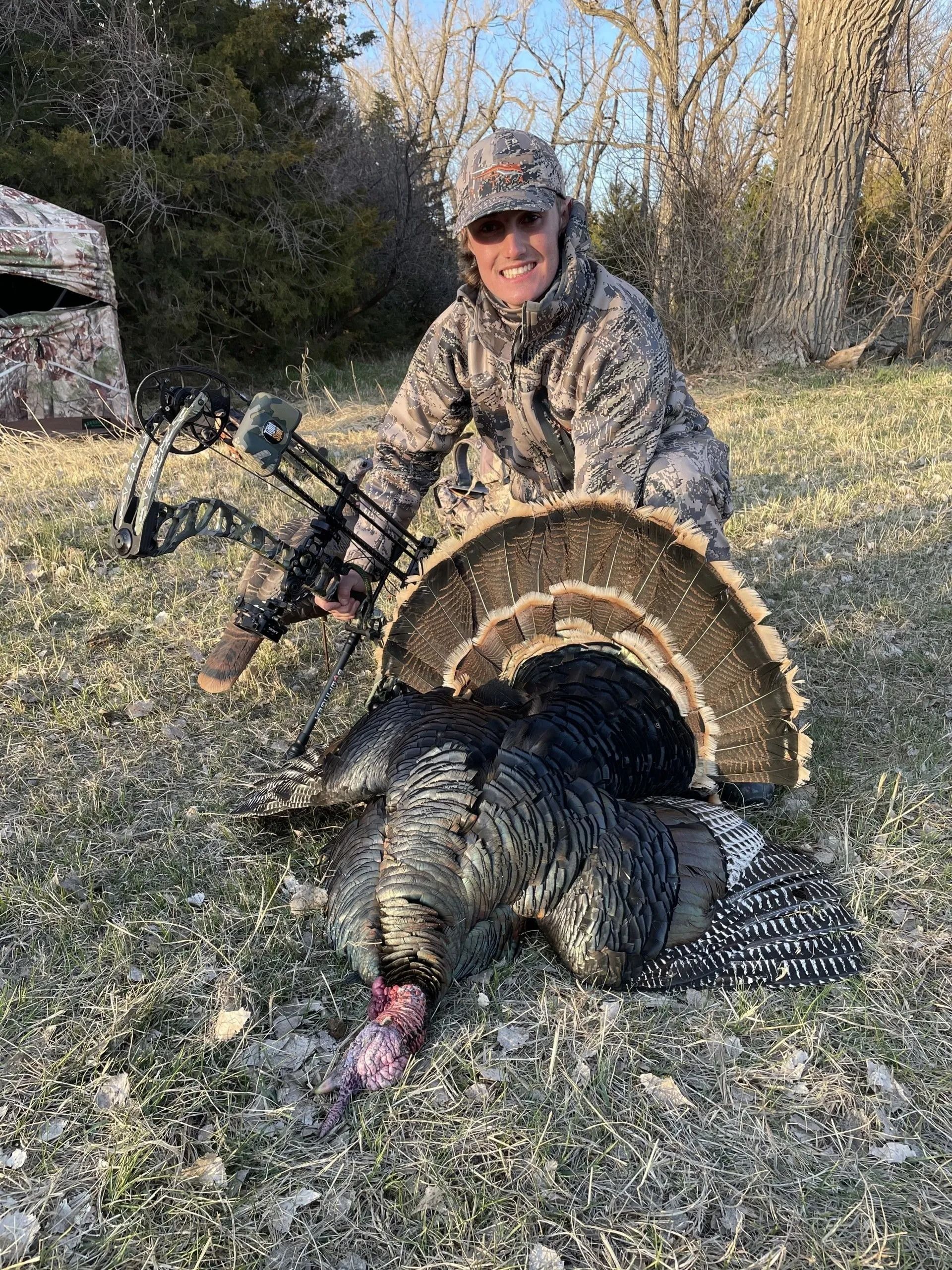 Hunter kneeling behind a wild turkey in a grassy field, holding up the bird by its tail fan.