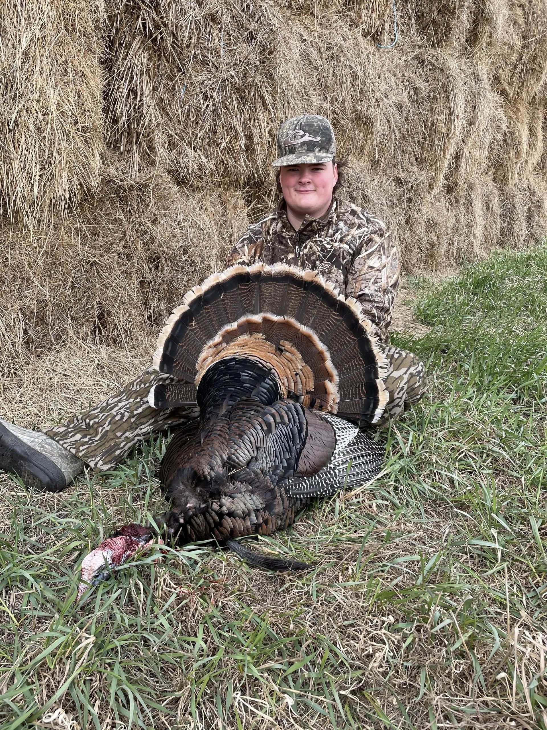 Hunter crouches behind a wild turkey with fanned tail in a grassy field against a hay bale backdrop