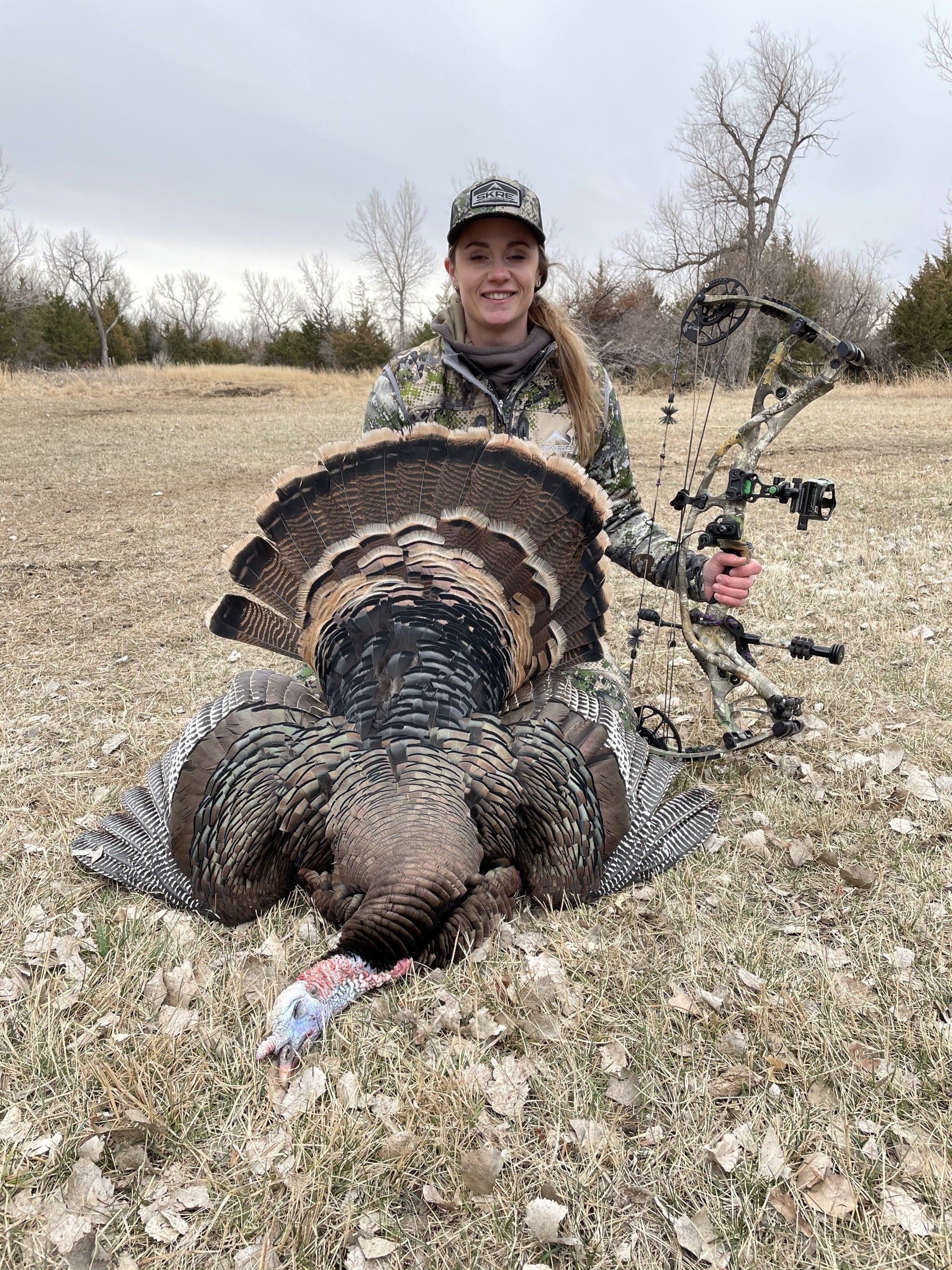 Hunter kneels behind a large wild turkey in a grassy field, holding its fan tail open.