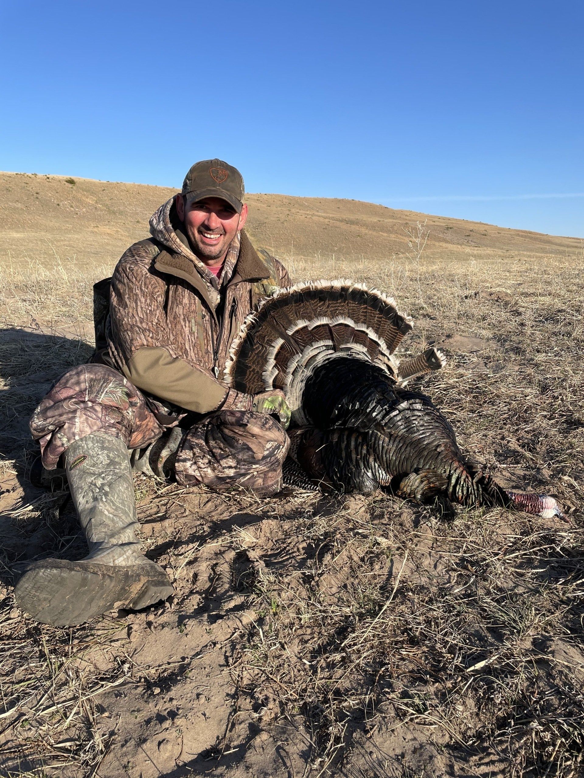 Hunter kneeling beside a large wild turkey in a dry, open field under a clear blue sky