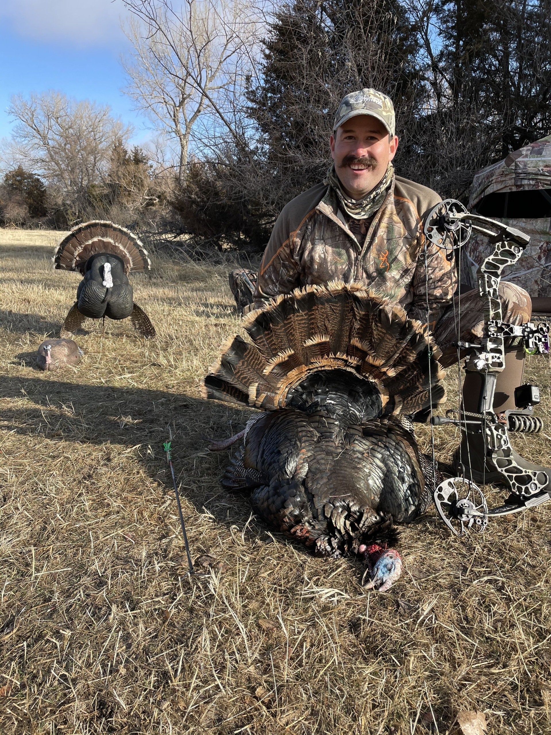 Hunter kneels beside two wild turkeys in a grassy field on a sunny day.