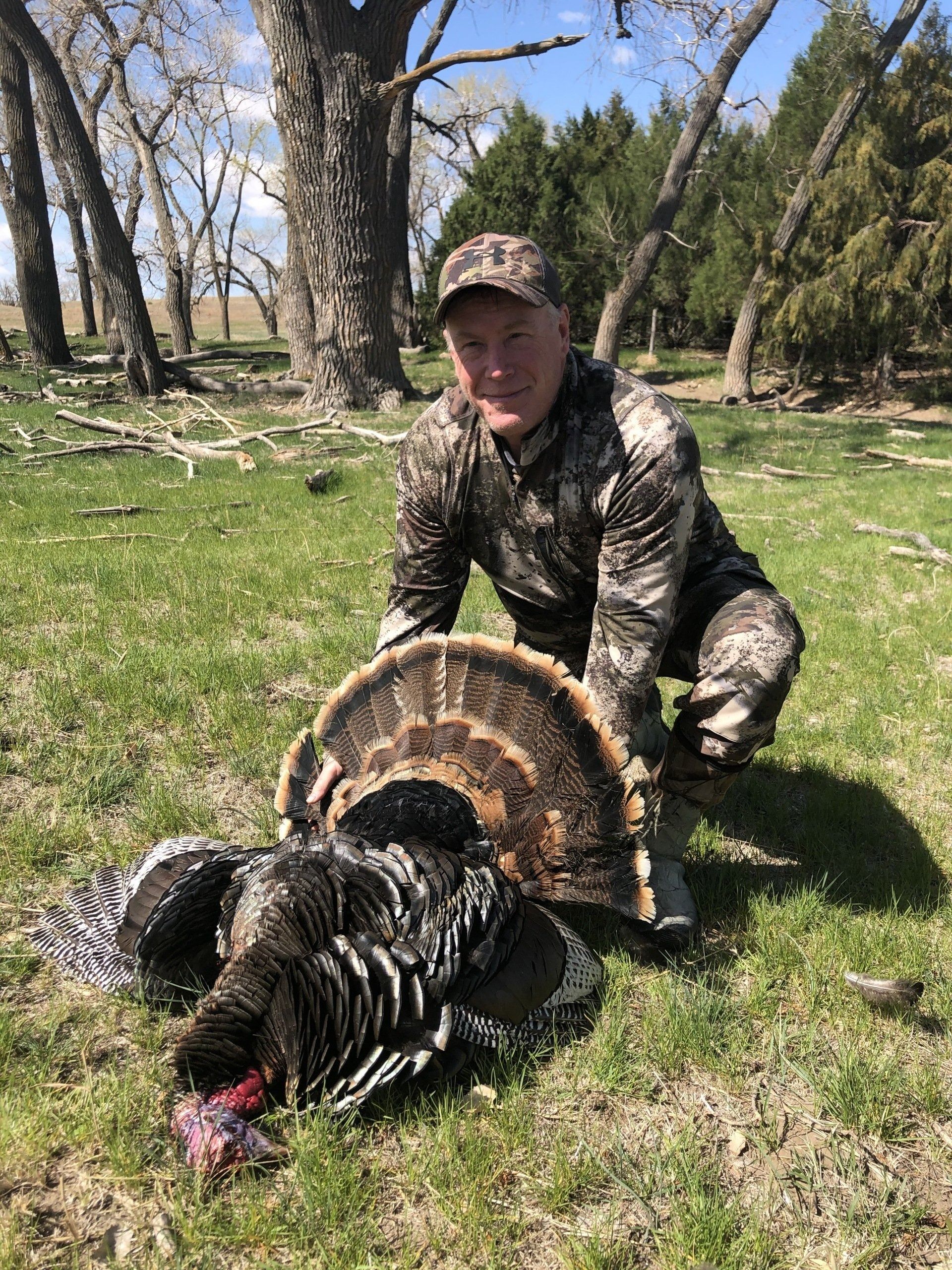Hunter kneeling beside a wild turkey in a wooded clearing on a sunny day