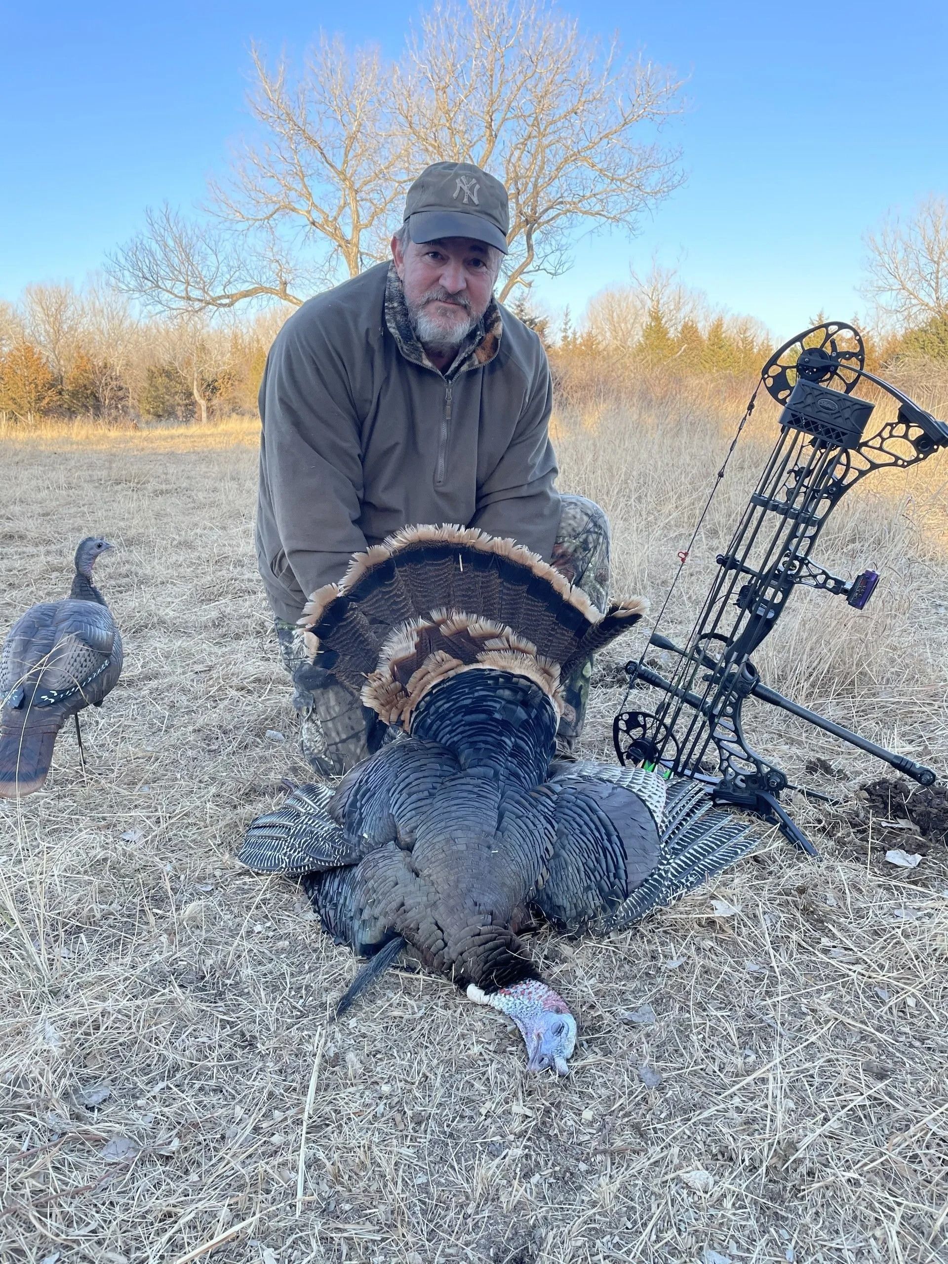 Hunter kneeling beside a large wild turkey in a grassy field with a compound bow nearby