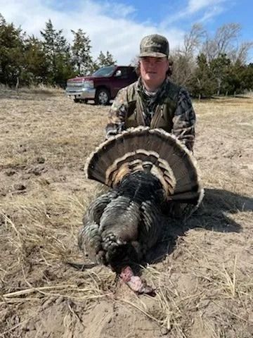 Hunter kneels behind a turkey he harvested in a grassy field, with a truck and trees in the background.