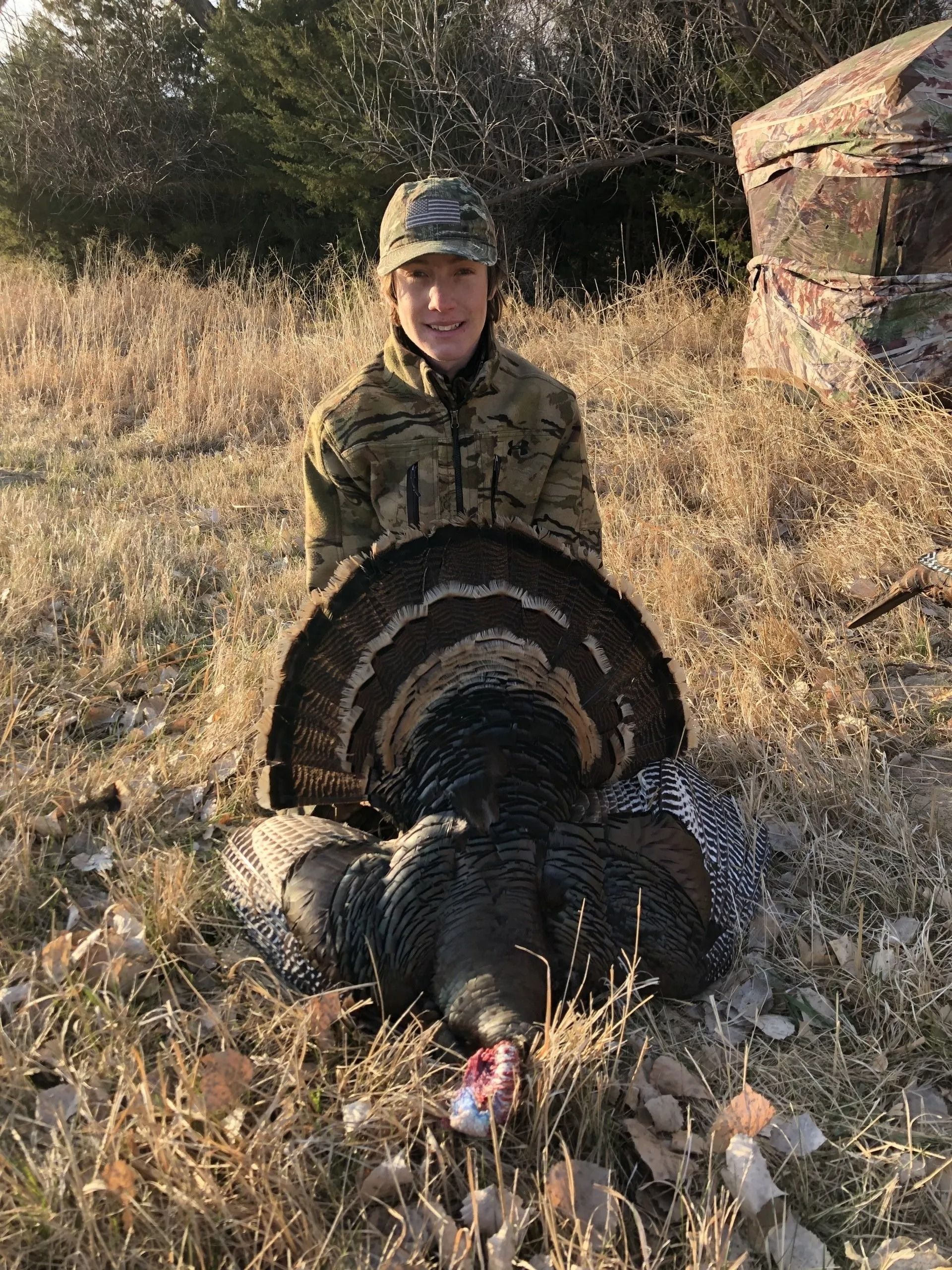 Hunter posing with a large turkey in tall grass beside rocks and trees.