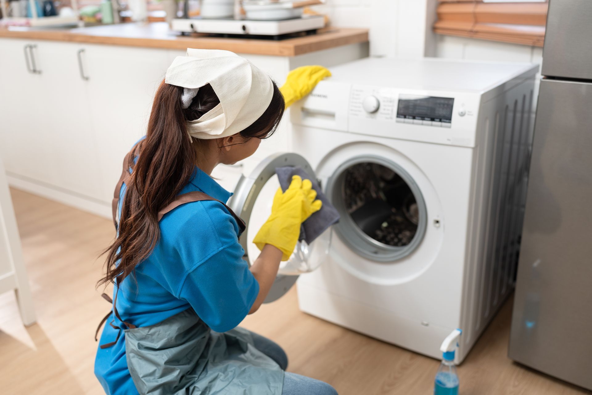 Person in blue cleaning a front-loading washing machine with yellow gloves in a laundry room