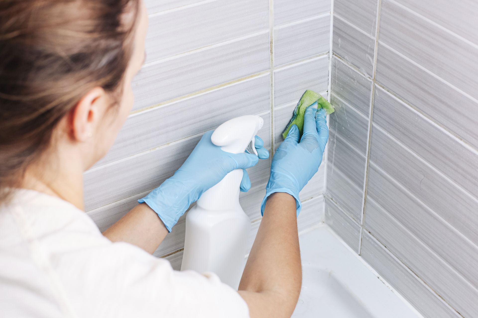 Person cleaning bathroom tiles with a spray bottle and cloth, wearing blue gloves