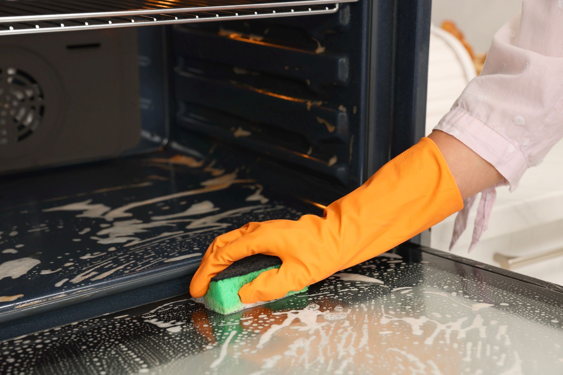 Gloved hand scrubbing the inside of a dirty oven with a green sponge