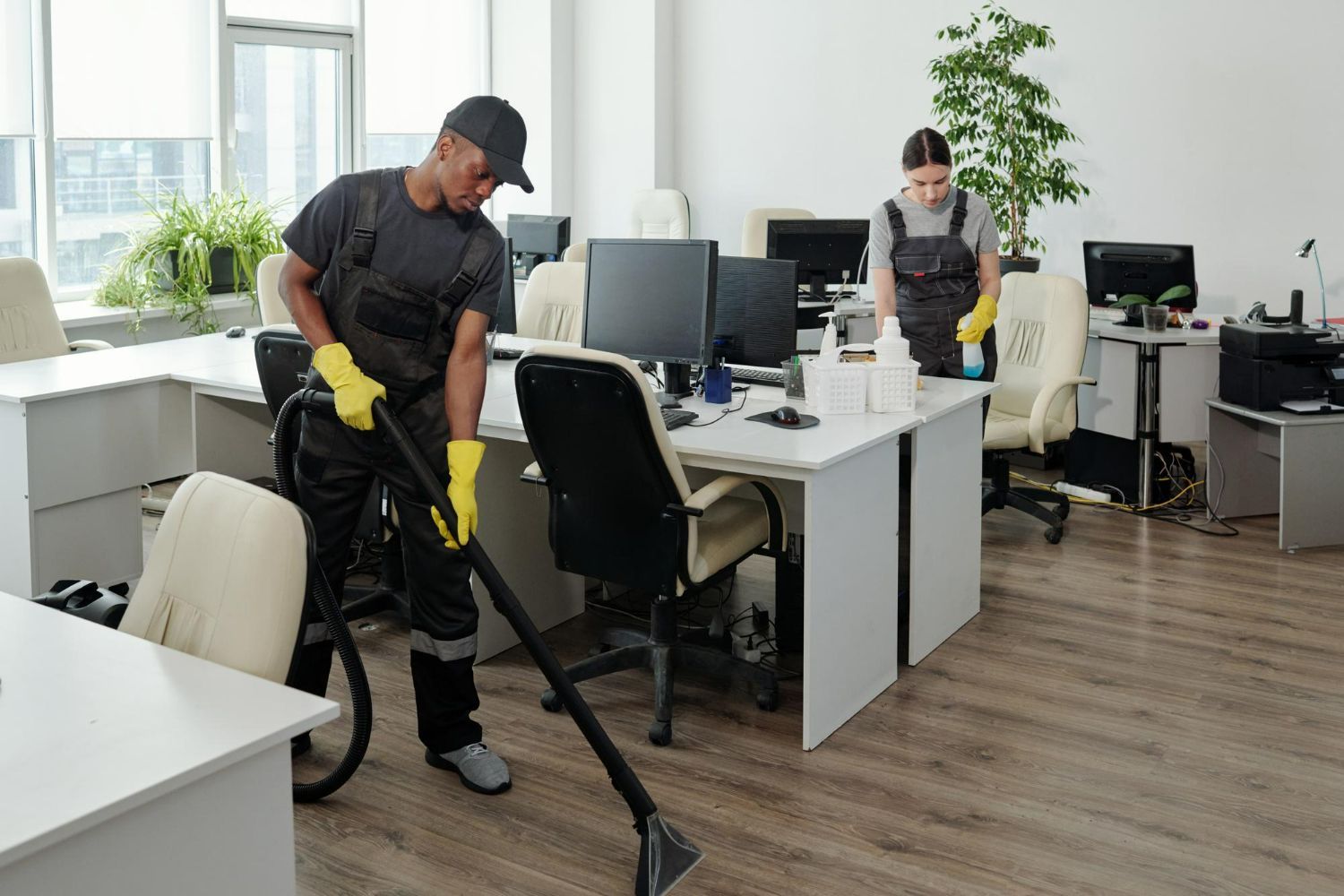 Two workers in grey uniforms and yellow gloves clean a modern office with desks, chairs, and computers.