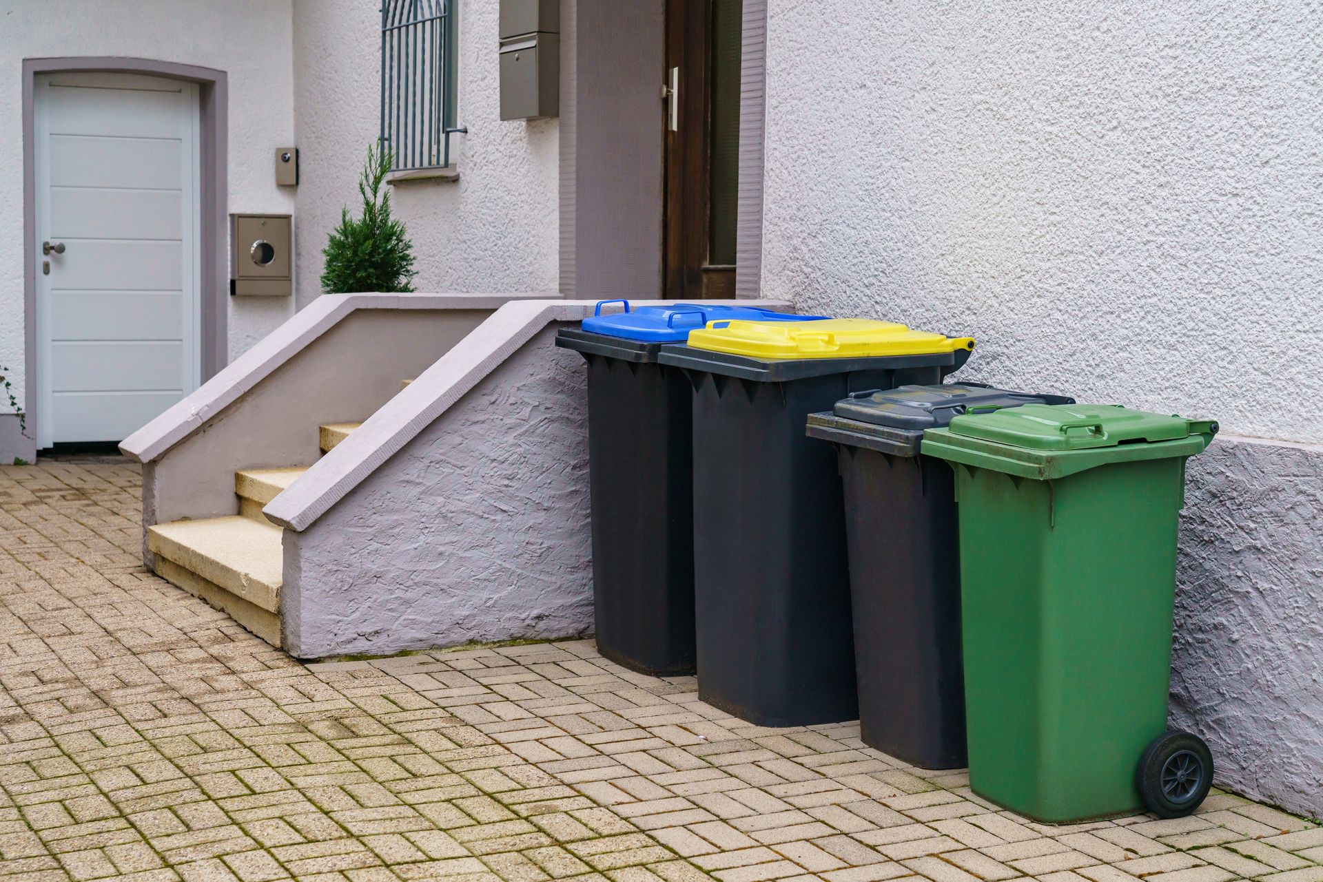 Trash bins lined up beside a house entrance on a cobblestone sidewalk.