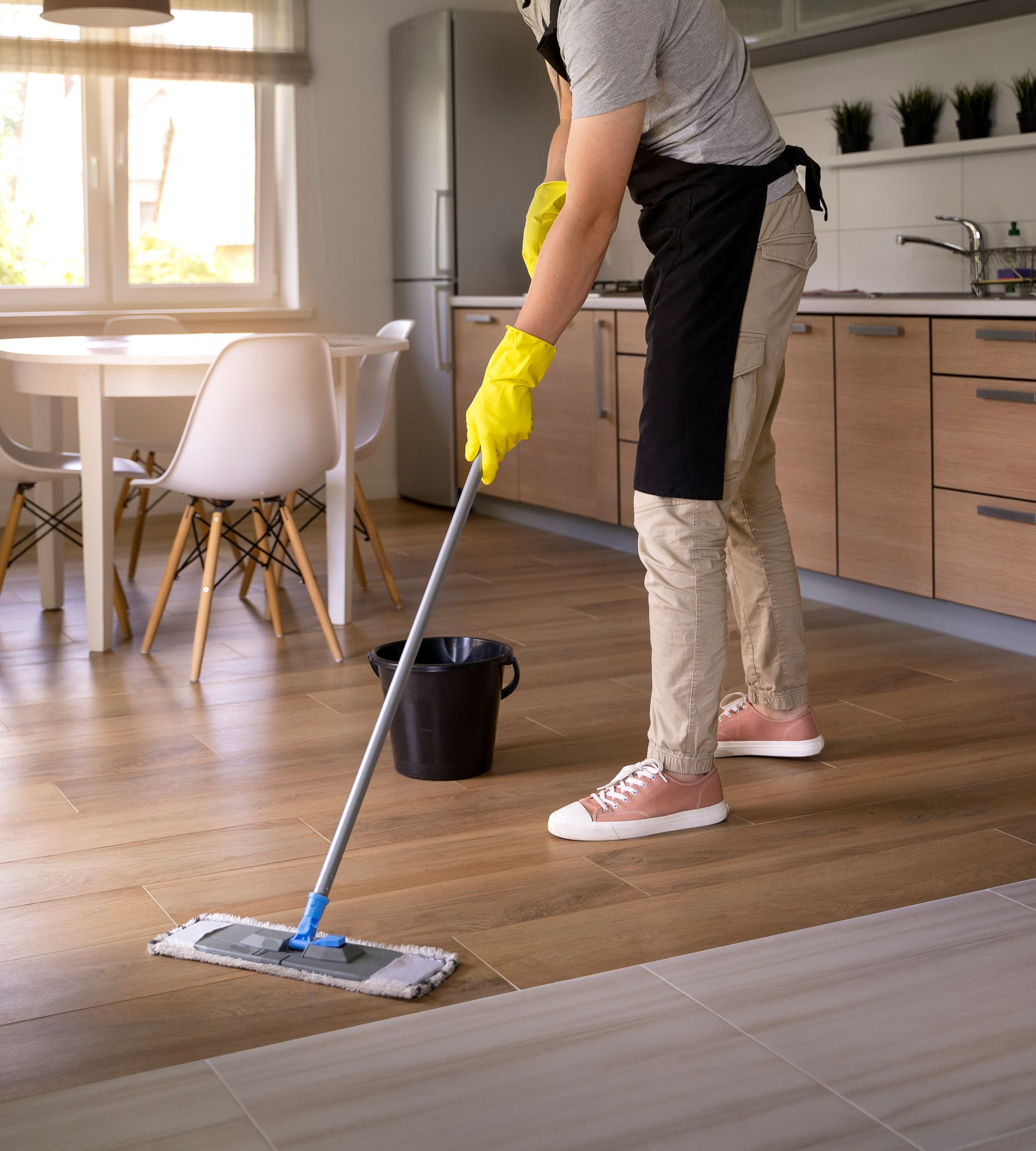 A person wearing a black apron and yellow gloves mops a wooden kitchen floor with a flat mop next to a black bucket.