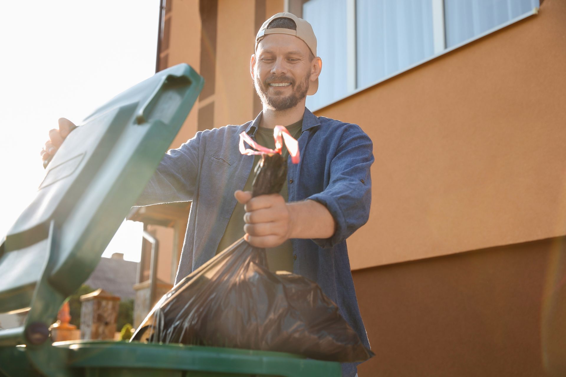 Man smiling, tossing trash bag into green bin outside a brown house.