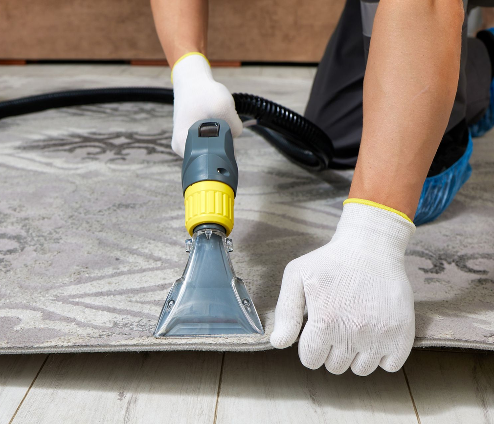 Person using a yellow-and-blue carpet cleaner to clean a gray rug on the floor.