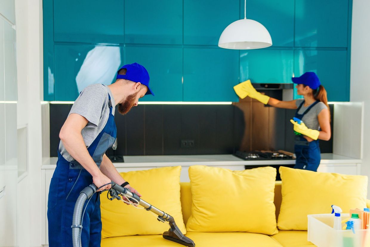 Two people cleaning a bright teal living room, one vacuuming a yellow sofa and the other wiping the wall.
