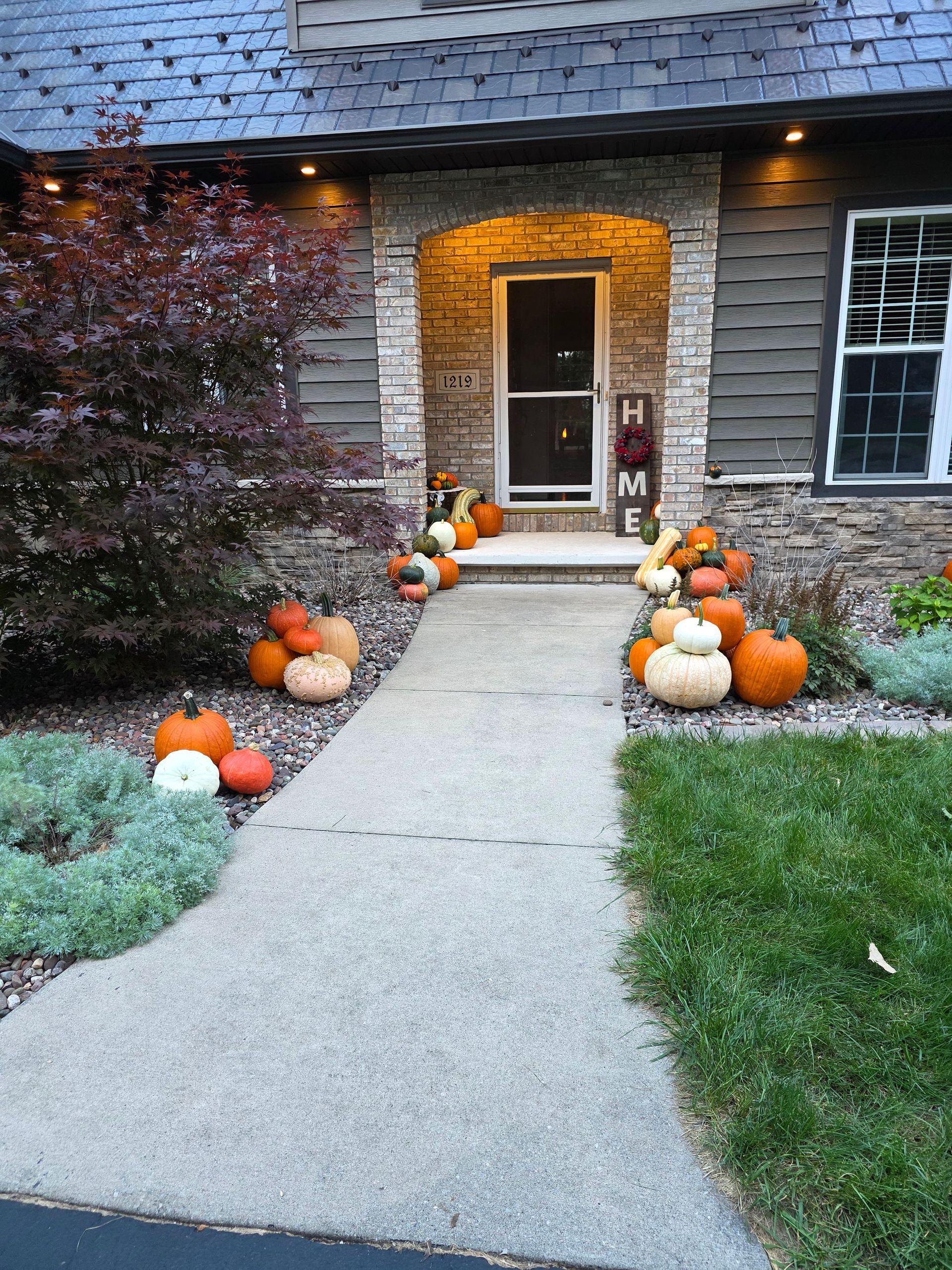 Pumpkins line a walkway to a front door decorated for fall, with a house featuring stone and gray siding.