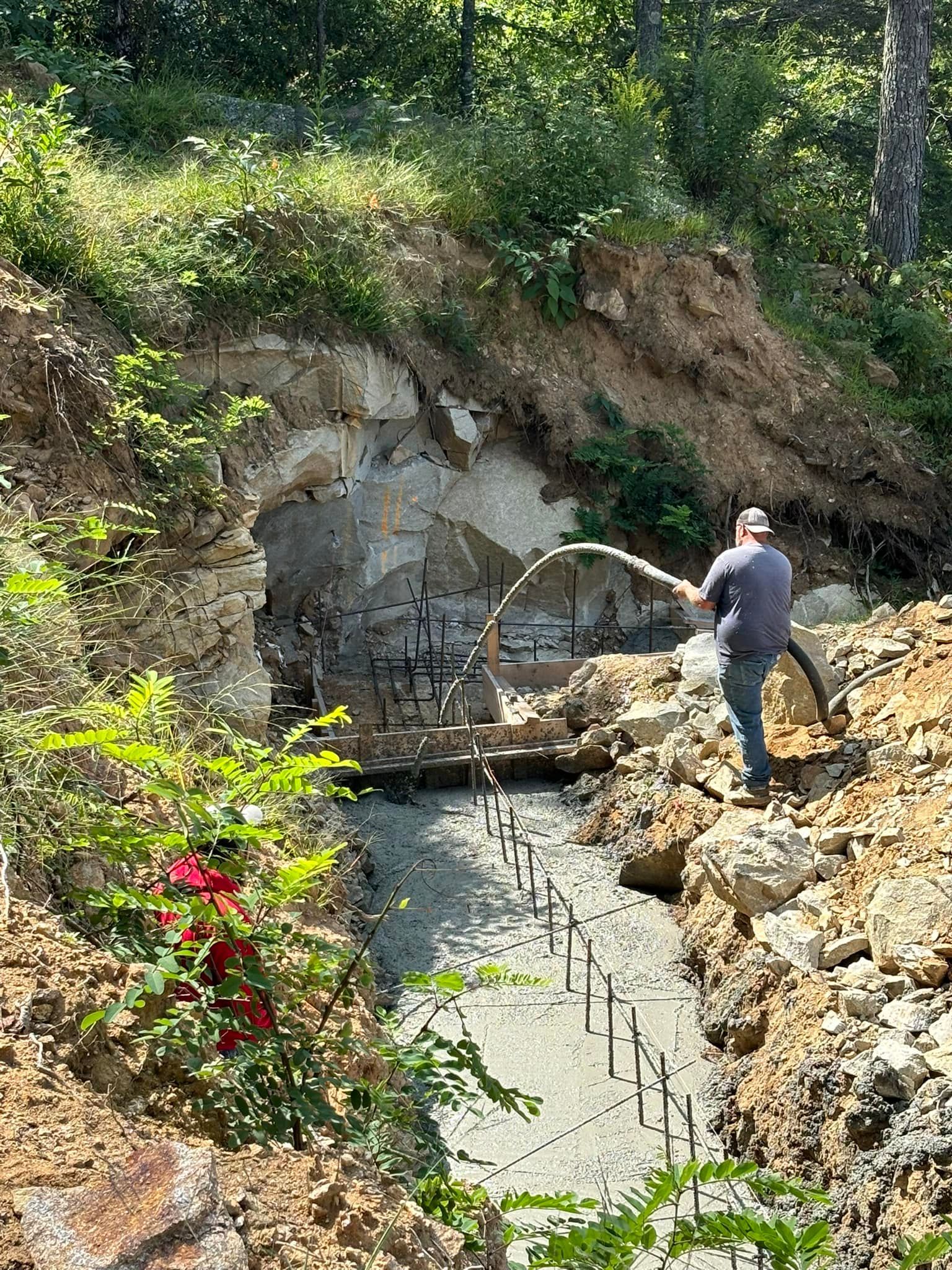 Construction worker spraying concrete into a cave-like structure; dirt and foliage surround.