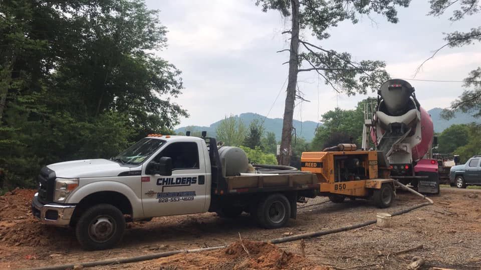 White truck towing concrete pump and cement truck on a dirt road, trees and mountains in the background.