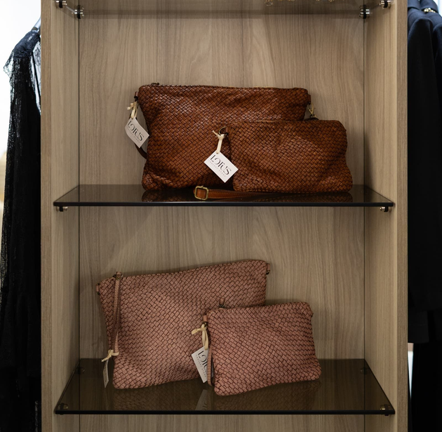 Una mano con le unghie rosa tocca una borsa di pelle marrone appoggiata su una superficie di legno.
