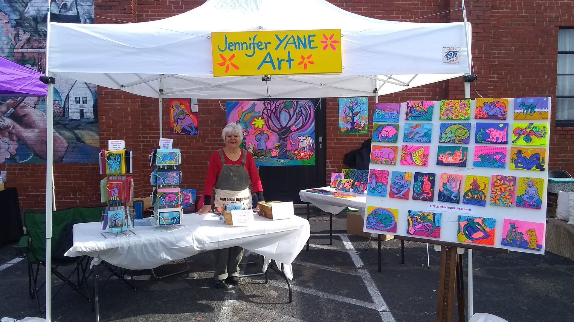 Artist Jennifer Yane stands at her art booth, displaying colorful paintings under a white tent.